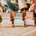 Three versions of a gold sled ornament stacked with pastel gift box with gold ribbon accents, displayed on a rustic wood slice with holiday decorations in the background.