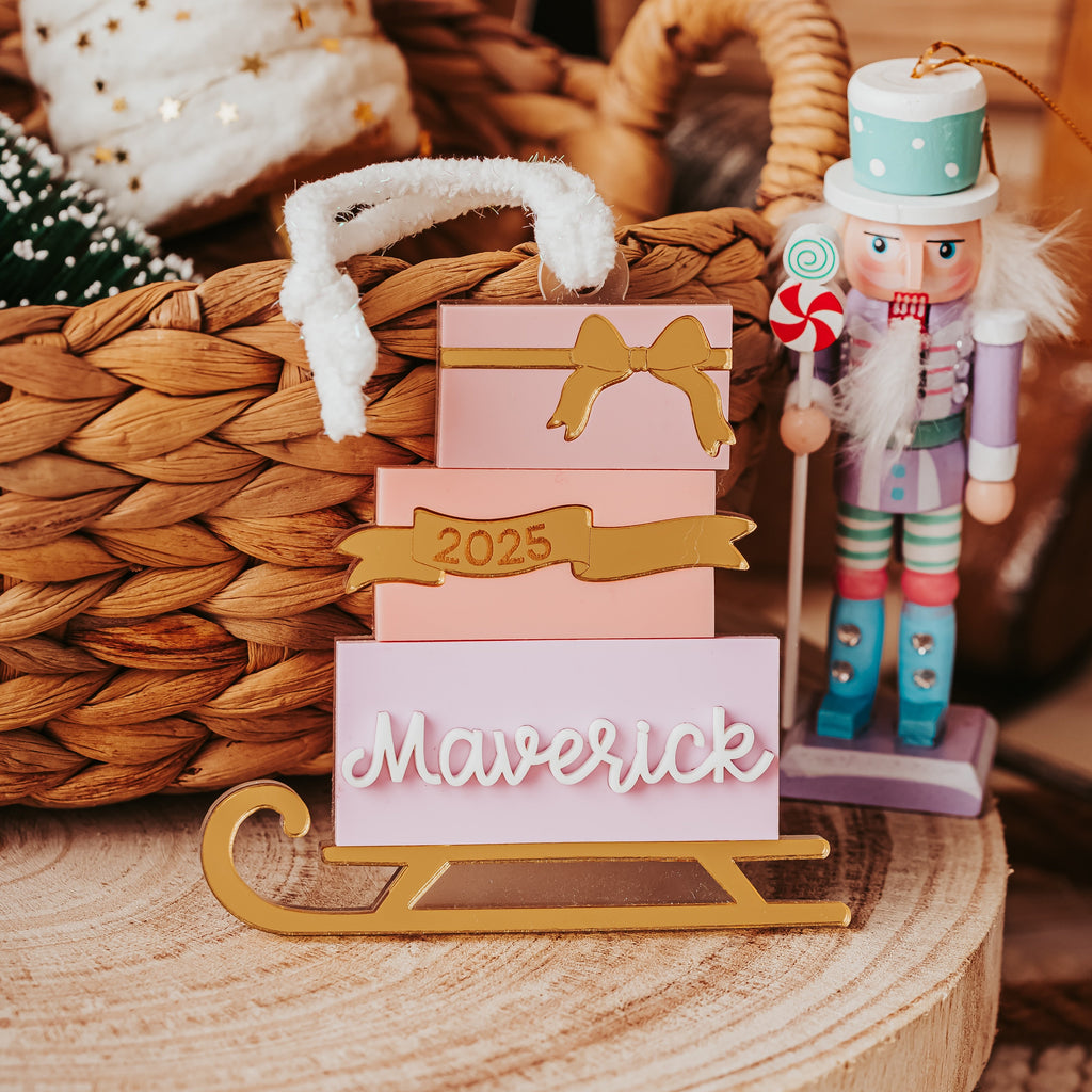 A personalized Christmas ornament display featuring various shades of pink gift boxes stack with gold accent ribbons, the year '2025', and the name 'Maverick' in white script on a gold sled base. A nutcracker stands to the right, and the scene is styled with a woven basket and holiday decor in warm, rustic tones.