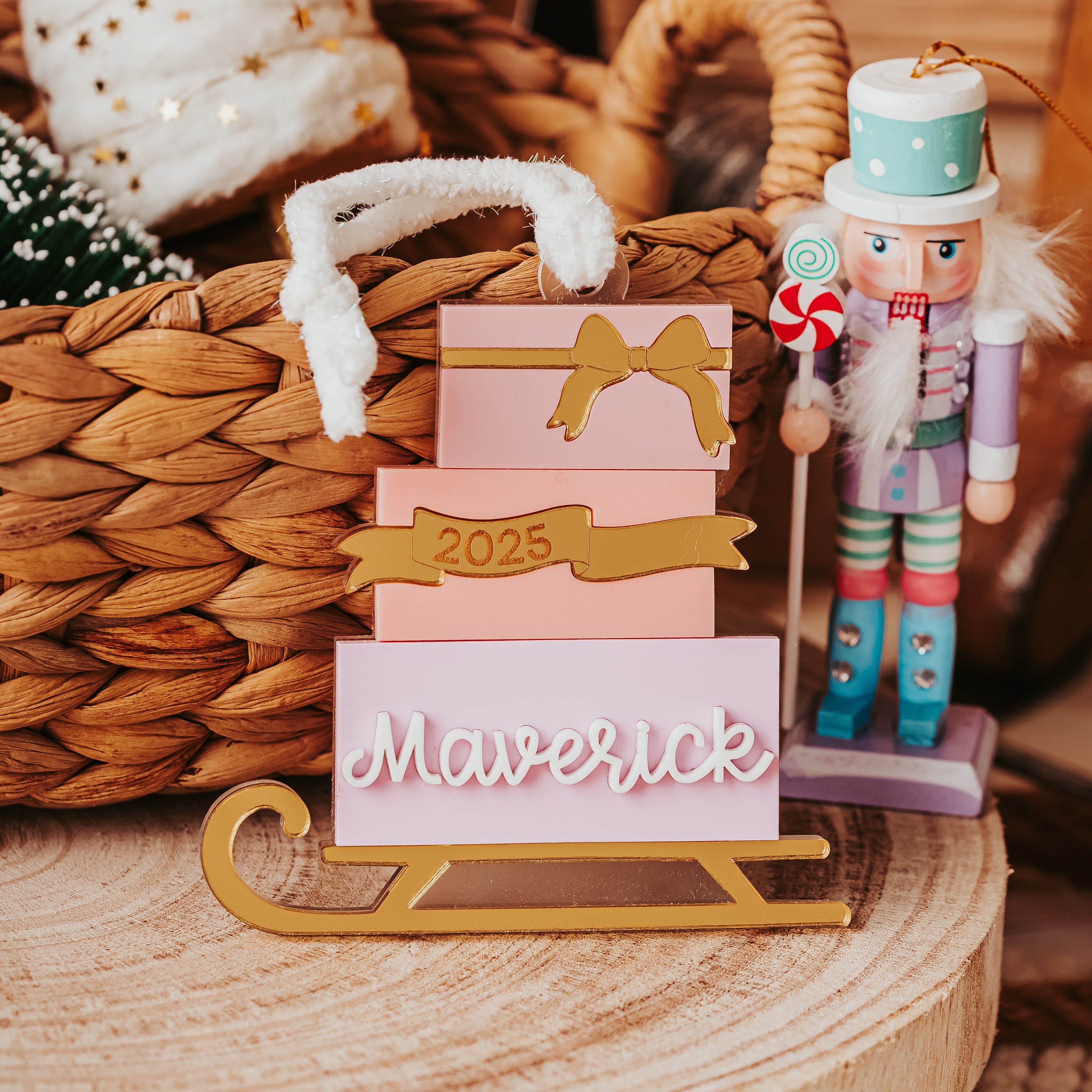 A personalized Christmas ornament display featuring various shades of pink gift boxes stack with gold accent ribbons, the year '2025', and the name 'Maverick' in white script on a gold sled base. A nutcracker stands to the right, and the scene is styled with a woven basket and holiday decor in warm, rustic tones.