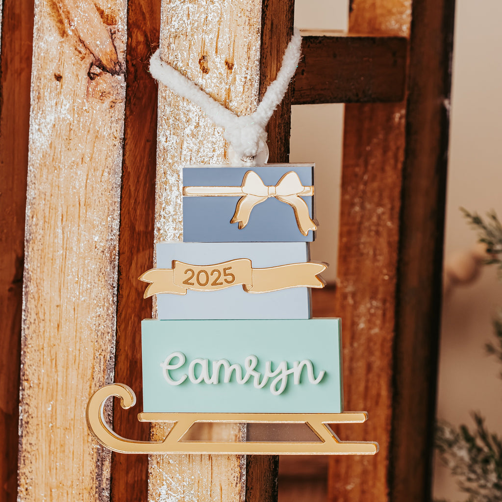 A personalized Christmas ornament shaped like a stack of gift boxes on a gold sled base , hanging against a rustic wooden background. The ornament features three stacked boxes in light blue, cream, and mint green, with a gold ribbon banner reading '2025' and a mint green box at the bottom with the name 'camryn' in white script.
