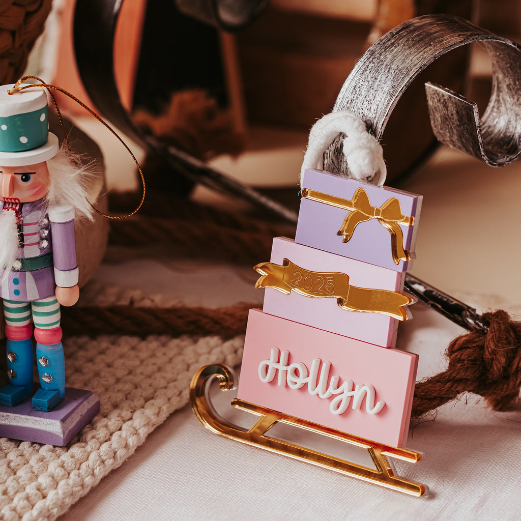 A close-up of a pink gift box stacked sled  ornament decorated with pastel purple gift boxes and gold ribbon details, alongside a nutcracker and rustic holiday decor.