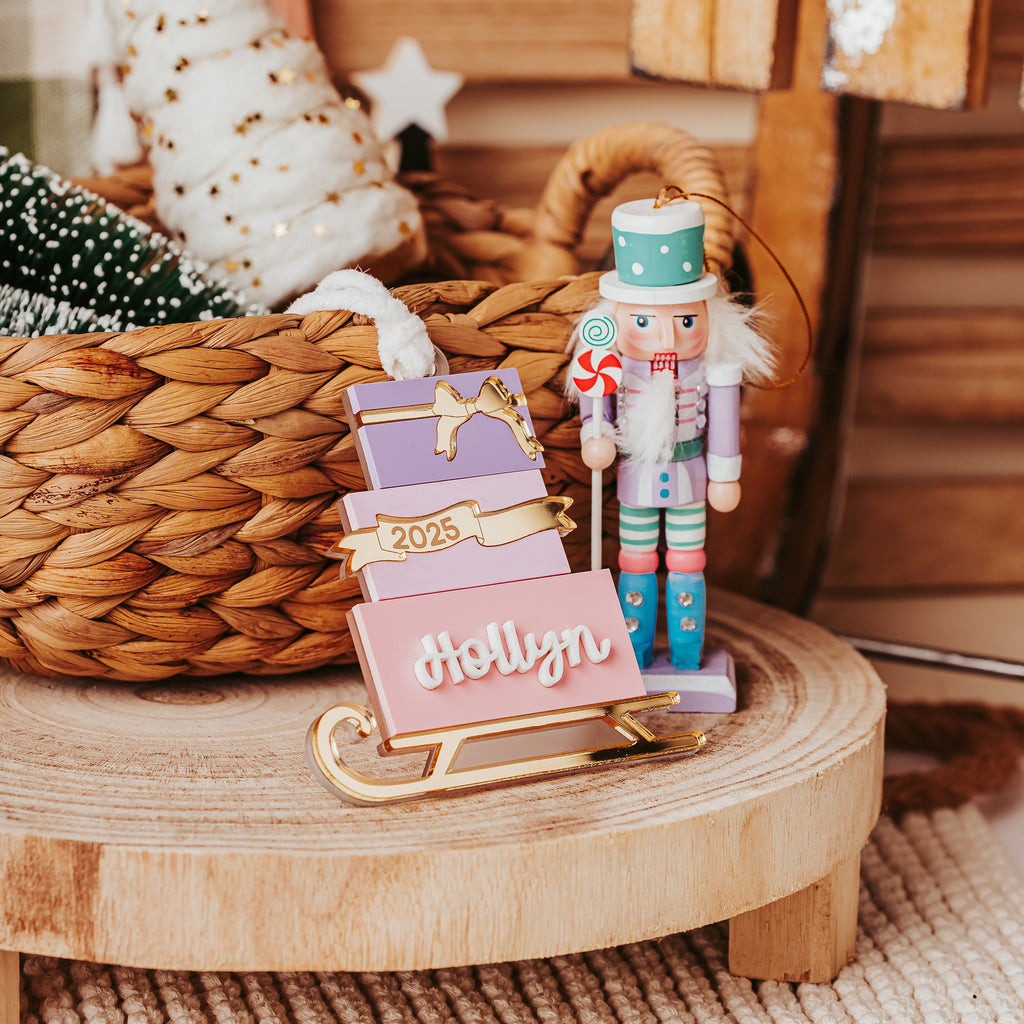 A personalized Christmas gift box stacked sled ornament  with the name 'Hollyn' and the year '2025', styled on a wooden tray with holiday decorations featuring a wooden nutcracker and felt trees.