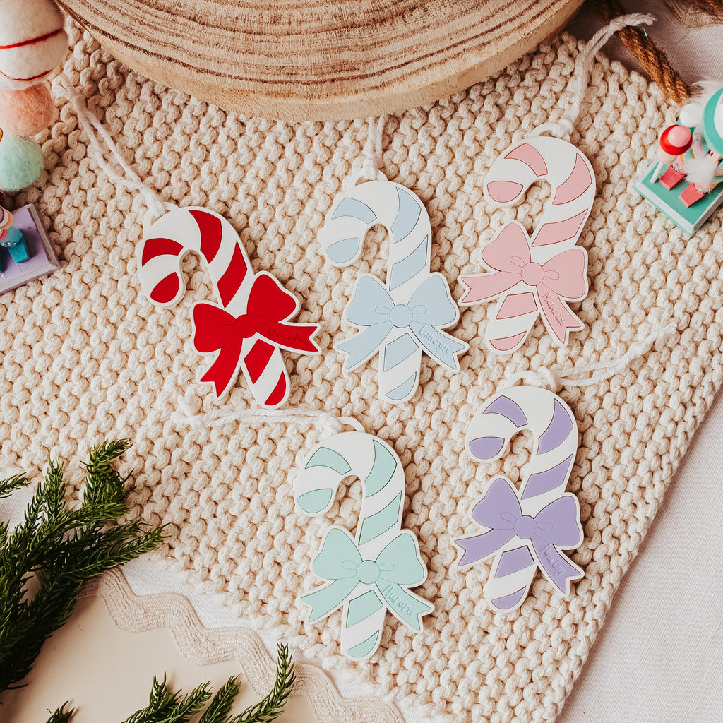 A styled flat-lay of six pastel-colored acrylic candy cane ornaments with bows, displayed on a beige knit blanket with holiday-themed props.