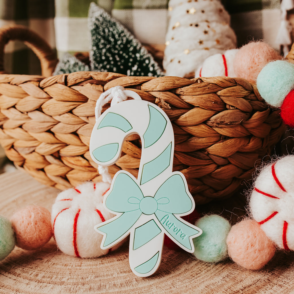 A personalized Christmas ornament shaped like a candy cane with a mint green bow and the name 'Aurora' in script, displayed in front of a woven basket with holiday decorations.