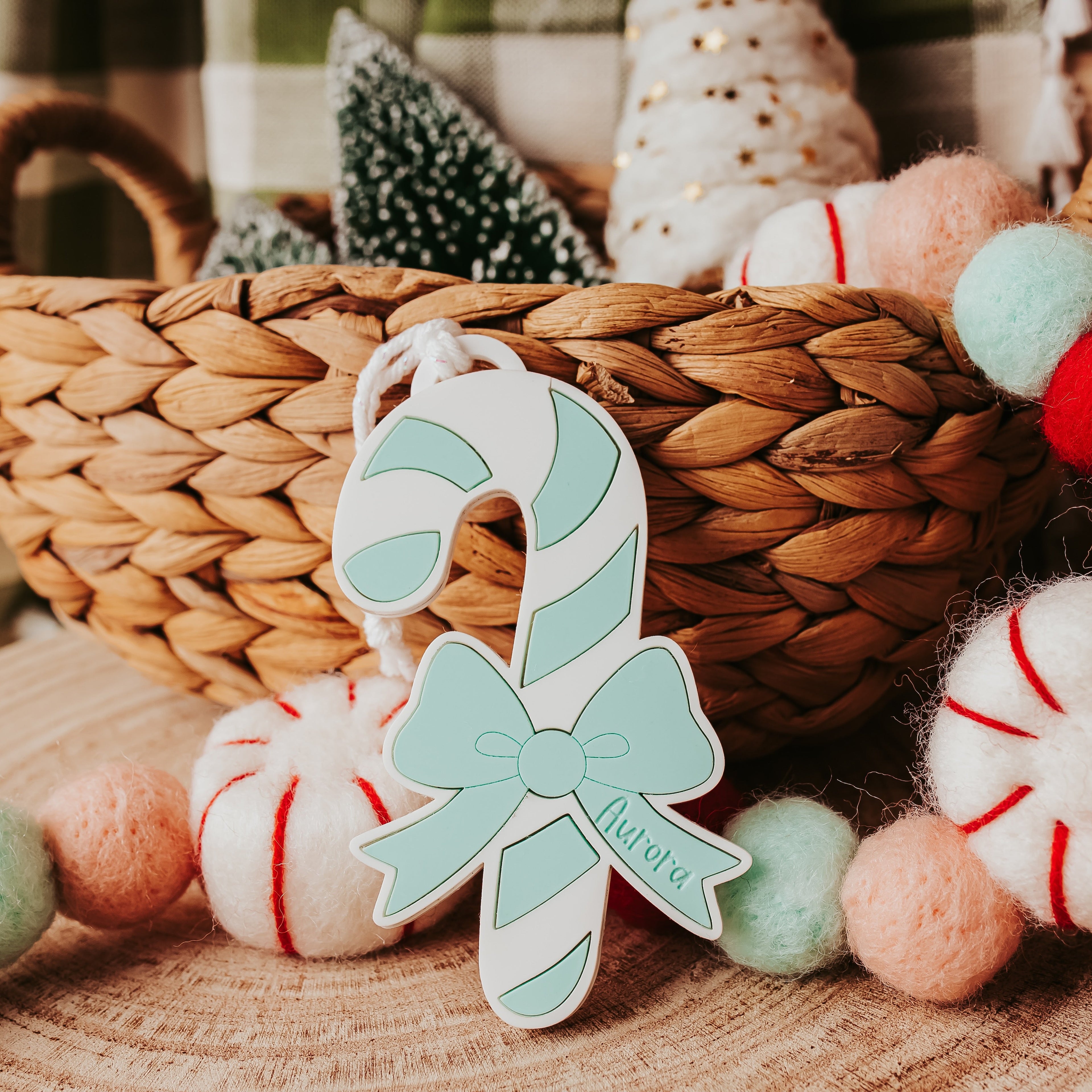 A personalized Christmas ornament shaped like a candy cane with a mint green bow and the name 'Aurora' in script, displayed in front of a woven basket with holiday decorations.
