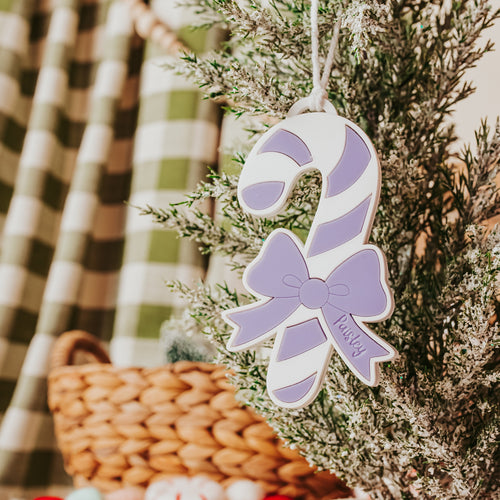 A personalized holiday ornament shaped like a candy cane with a bow, hanging on a small frosted green tree. The ornament is white with lavender purple stripes and a bow, featuring the name 'Paisley' in script.