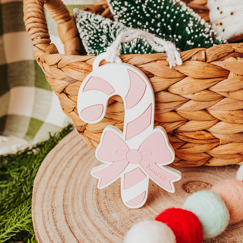 A pink and white candy cane ornament with a bow and the name 'Maverick' printed on it, hanging in a woven basket with Christmas decorations.