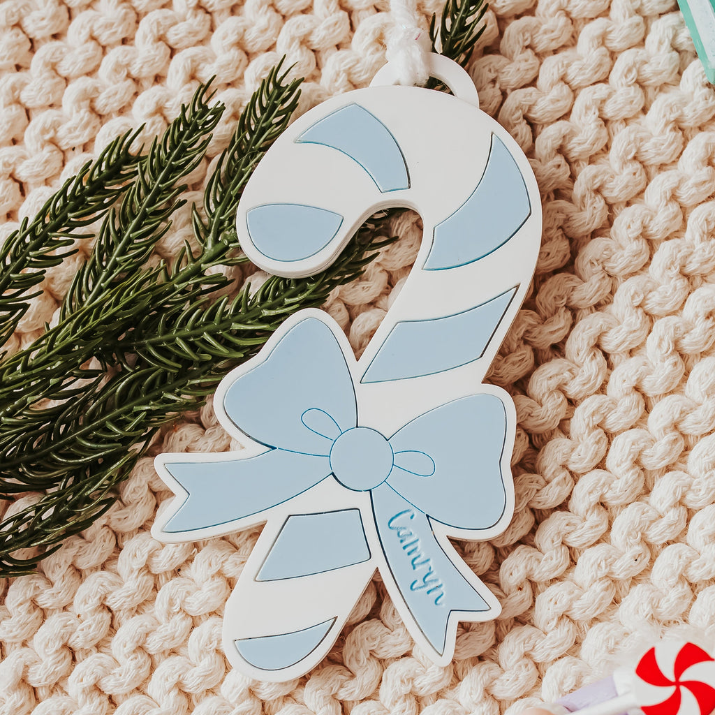 A light blue and white candy cane Christmas ornament with a bow and the name 'Camryn' written on it, styled on a beige knit blanket with green pine branches and a nutcracker toy.