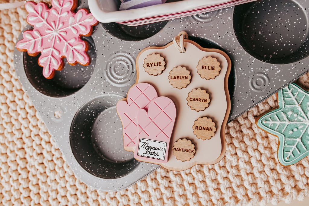 A styled flat-lay of a personalized baking oven mitt with cookie cutters, placed on a speckled gray baking tray over a beige knit blanket. The mitt is pink and beige with custom names printed on cookie-shaped cutters.