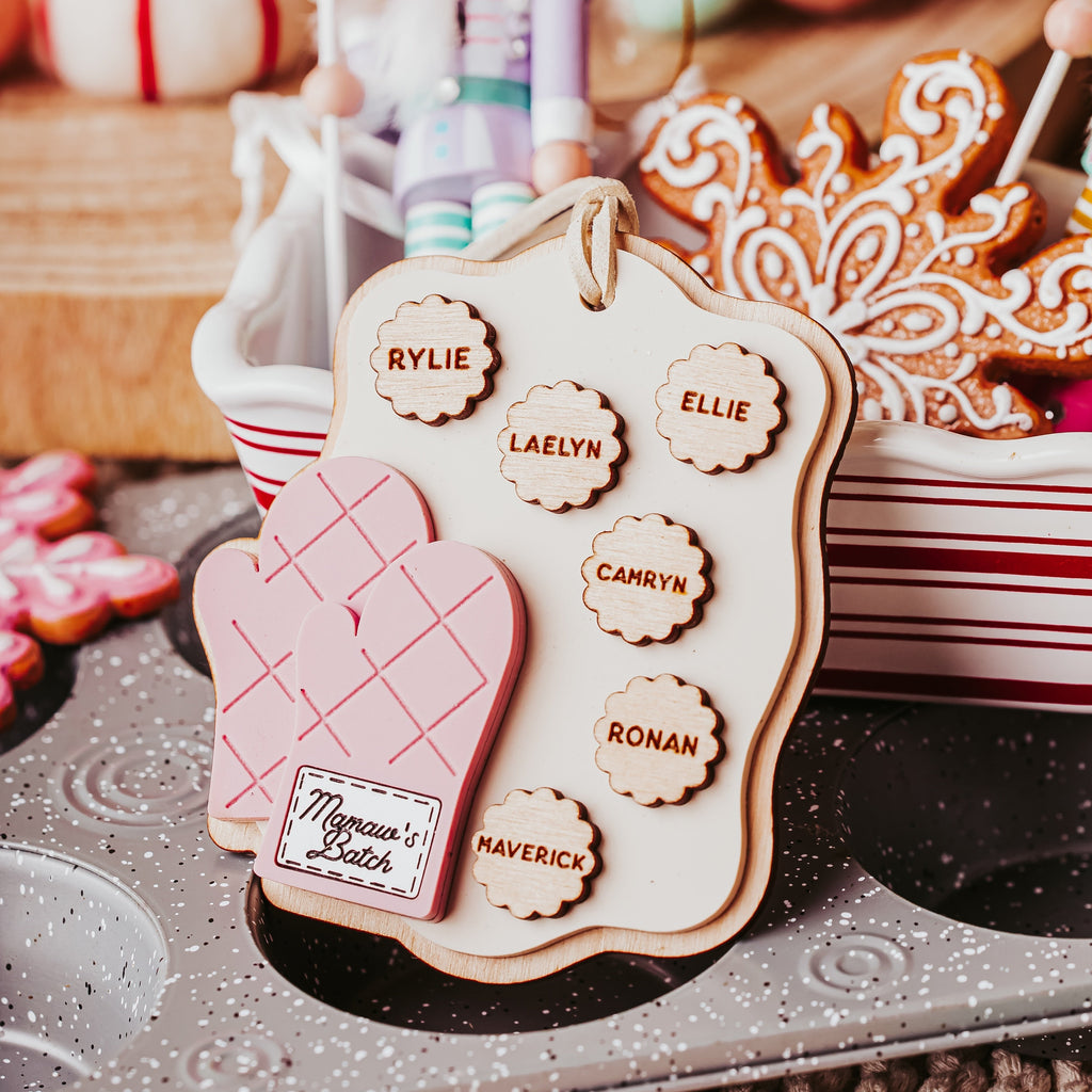 A close-up holiday scene showing a gingerbread cookie ornament shaped like a pink oven mitt with a name tag reading 'Mamaw's Batch,' alongside other decorated cookies and a nutcracker figurine in a festive tabletop setting.