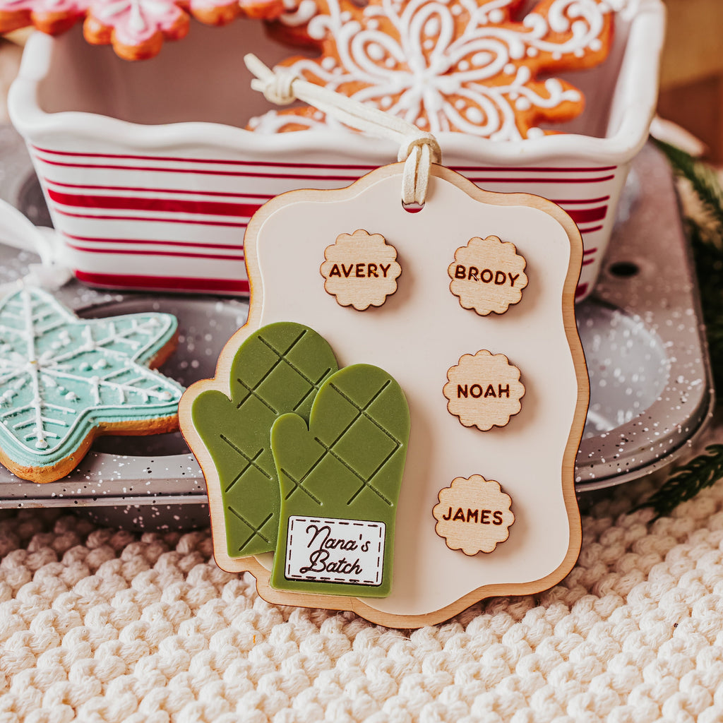 A personalized Christmas ornament shaped like a cookie tray with green mittens and a hanging loop, displayed on a tray with gingerbread cookies and holiday decorations in the background.