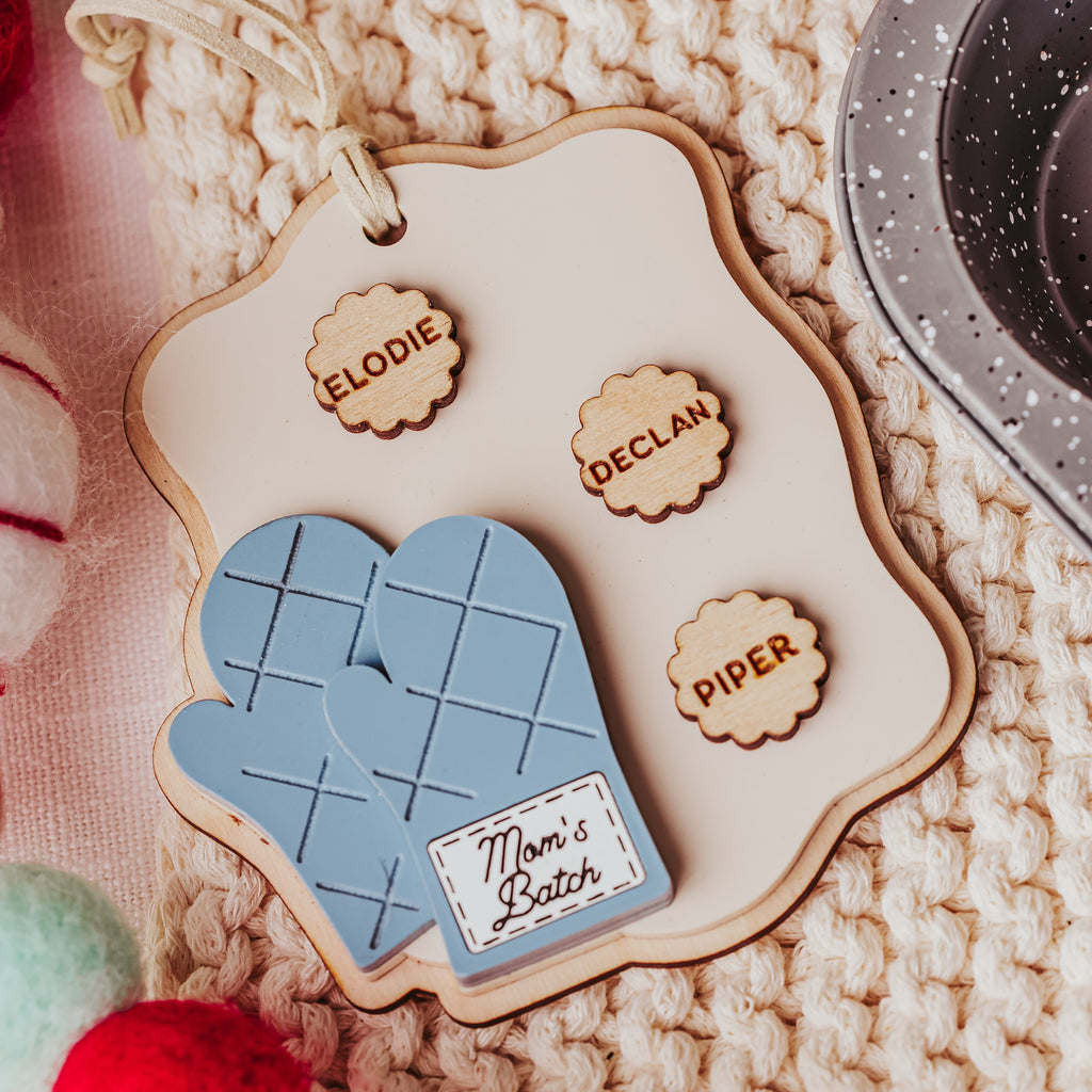 A personalized wooden oven mitt ornament featuring a light blue quilted mitt design, a cream-colored wooden plaque with laser-engraved names, and a small wooden tag reading 'Mom's Batch'. The ornament is styled on a cozy beige chunky-knit blanket with festive red, white, and green pom-poms.