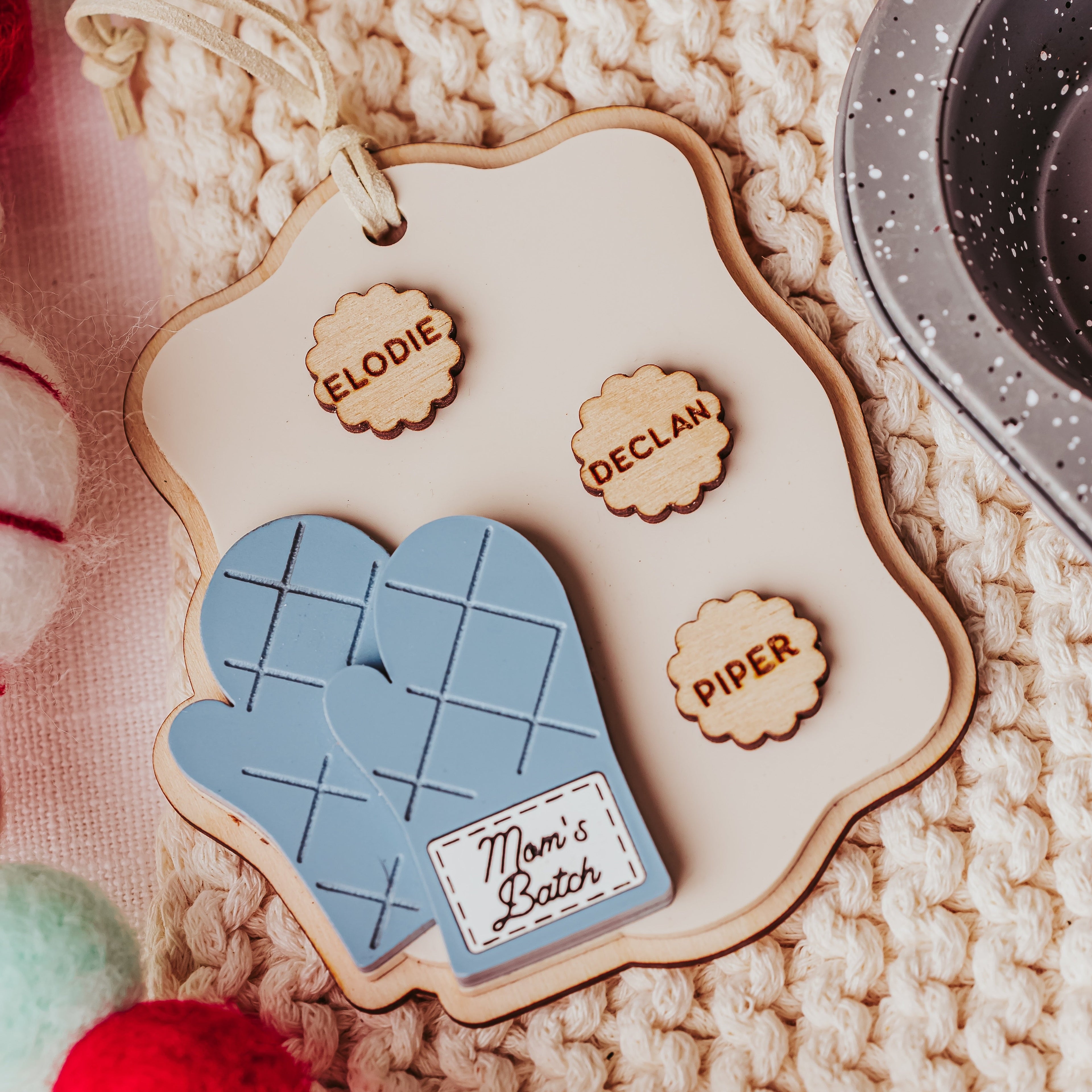 A personalized wooden oven mitt ornament featuring a light blue quilted mitt design, a cream-colored wooden plaque with laser-engraved names, and a small wooden tag reading 'Mom's Batch'. The ornament is styled on a cozy beige chunky-knit blanket with festive red, white, and green pom-poms.