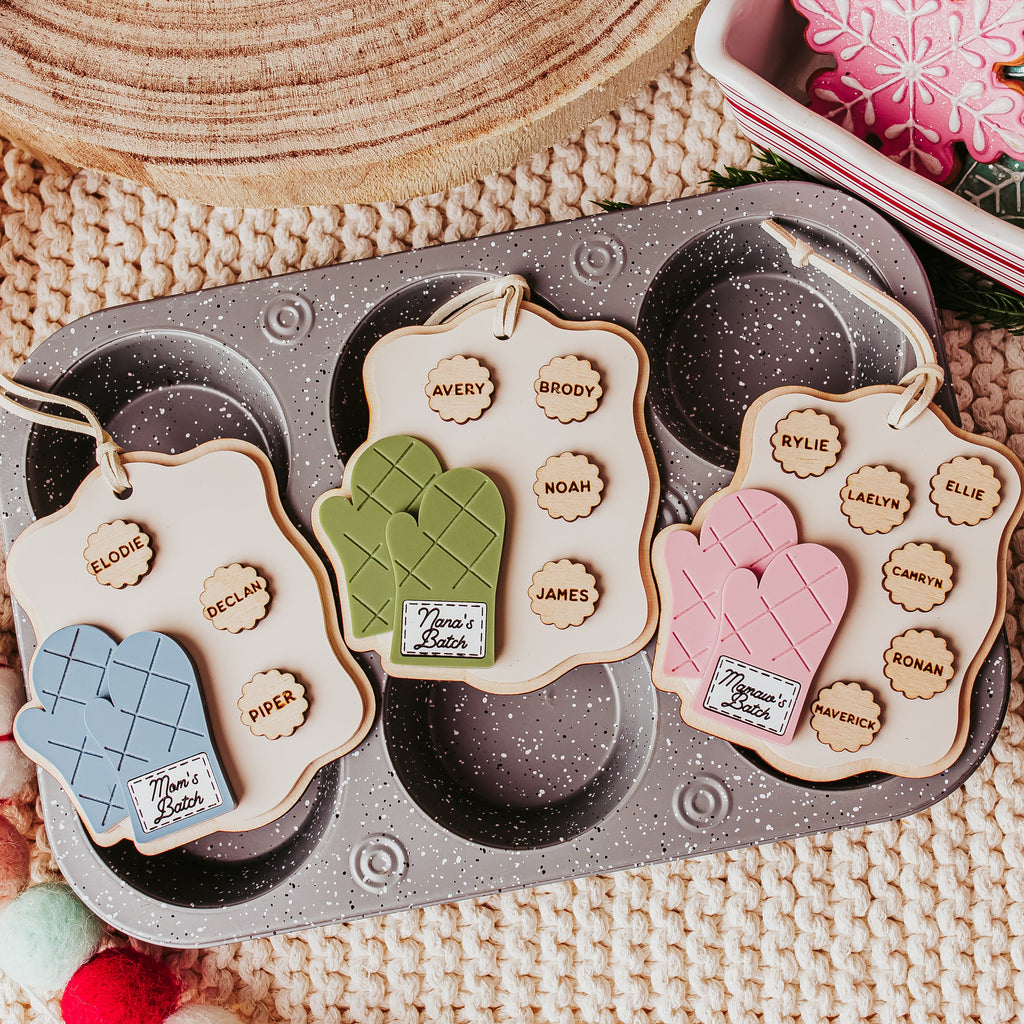 Three oven mitt-shaped Christmas ornaments with names on tags, displayed on a gray muffin tray over a beige knit blanket with holiday decorations.