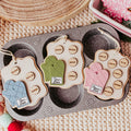 Three oven mitt-shaped Christmas ornaments with names on tags, displayed on a gray muffin tray over a beige knit blanket with holiday decorations.