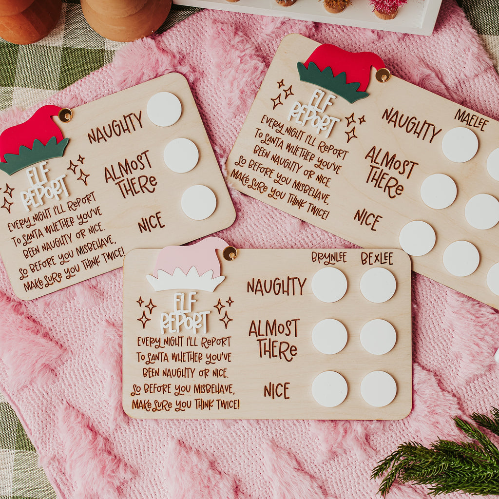 Three personalized Christmastelf report cards laid out on a pink blanket over a green-and-white checkered tablecloth, with pine branches and a dry erase marker nearby.