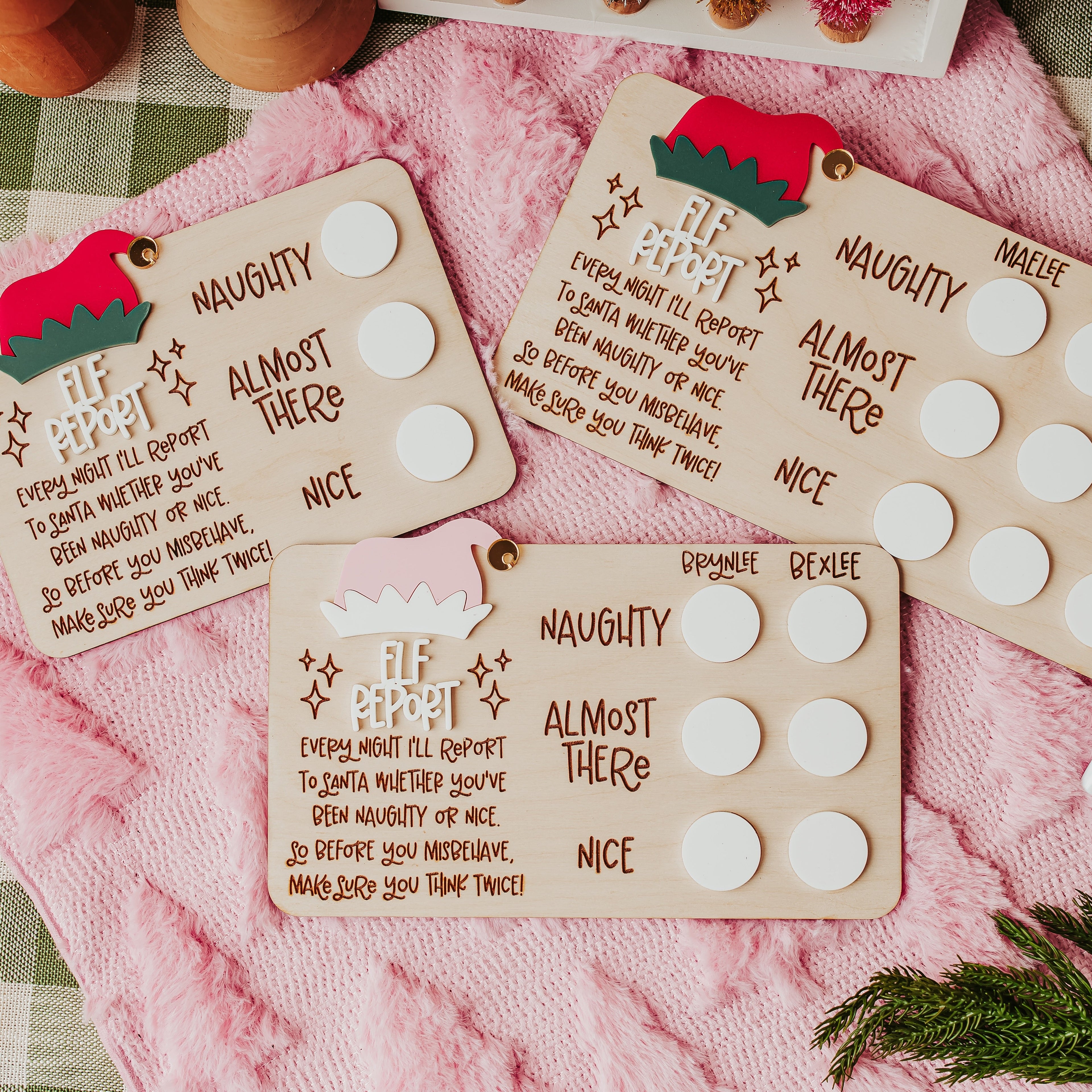 Three personalized Christmastelf report cards laid out on a pink blanket over a green-and-white checkered tablecloth, with pine branches and a dry erase marker nearby.