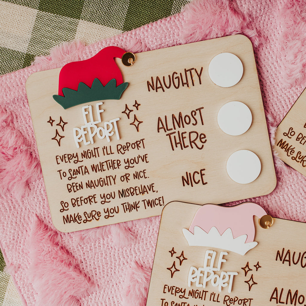 Three wooden Elf Report dry erase cards for naughty or nice behavior, decorated with festive red and pink hats and displayed on a pink fuzzy blanket with a plaid tablecloth and evergreen branches.