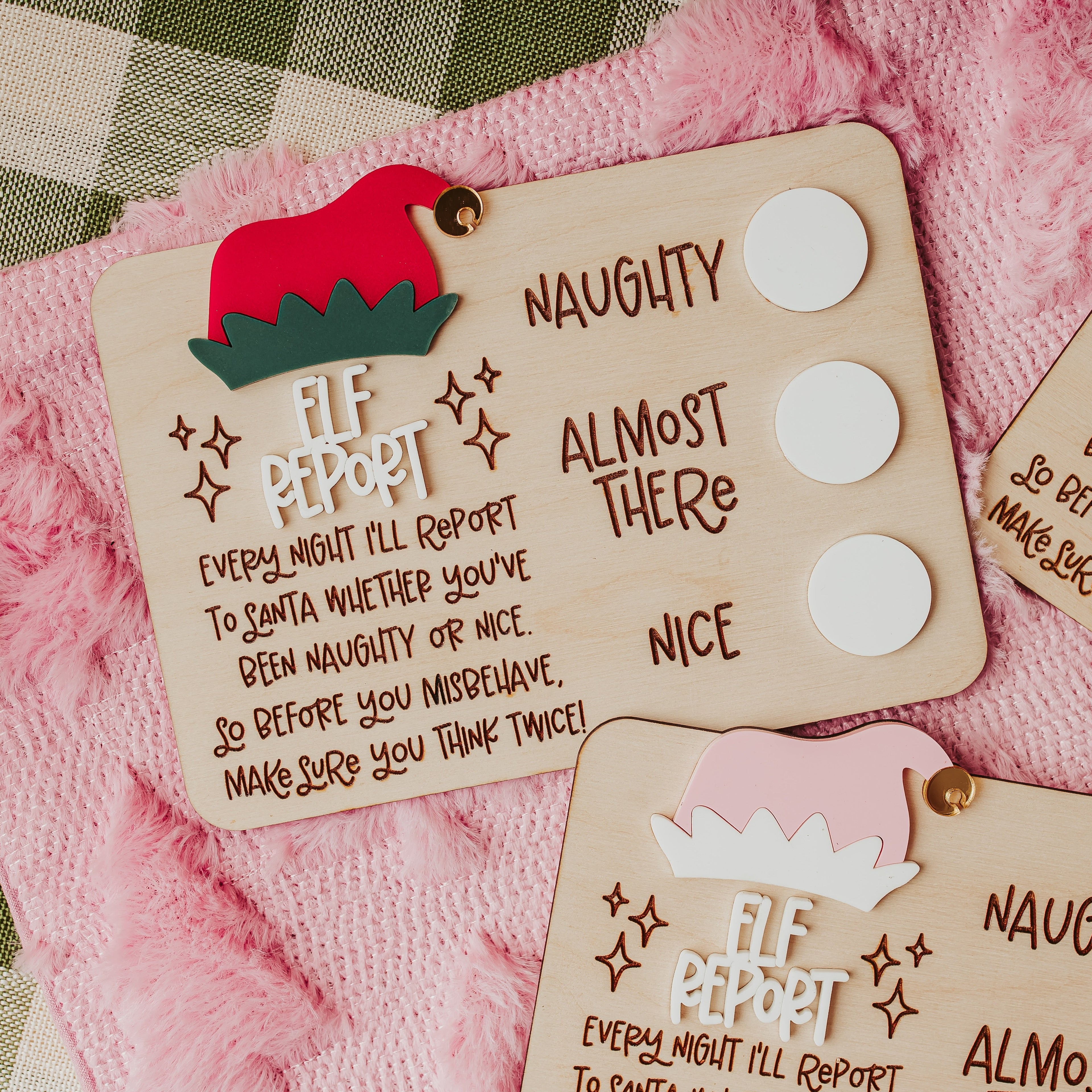 Three wooden Elf Report dry erase cards for naughty or nice behavior, decorated with festive red and pink hats and displayed on a pink fuzzy blanket with a plaid tablecloth and evergreen branches.
