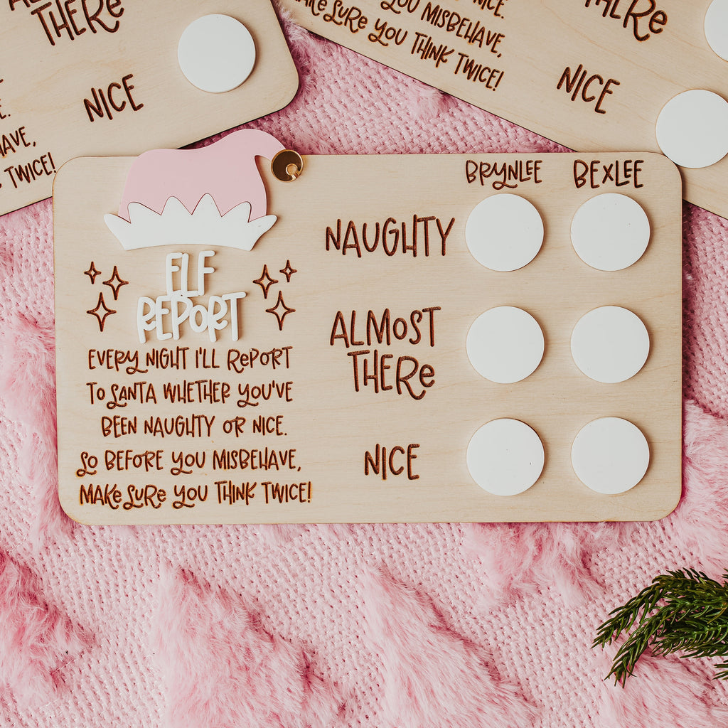 A close-up of a wooden Elf Report card with white circular white acrylic with personalized names, placed on a pink knit blanket with other matching cards partially visible.
