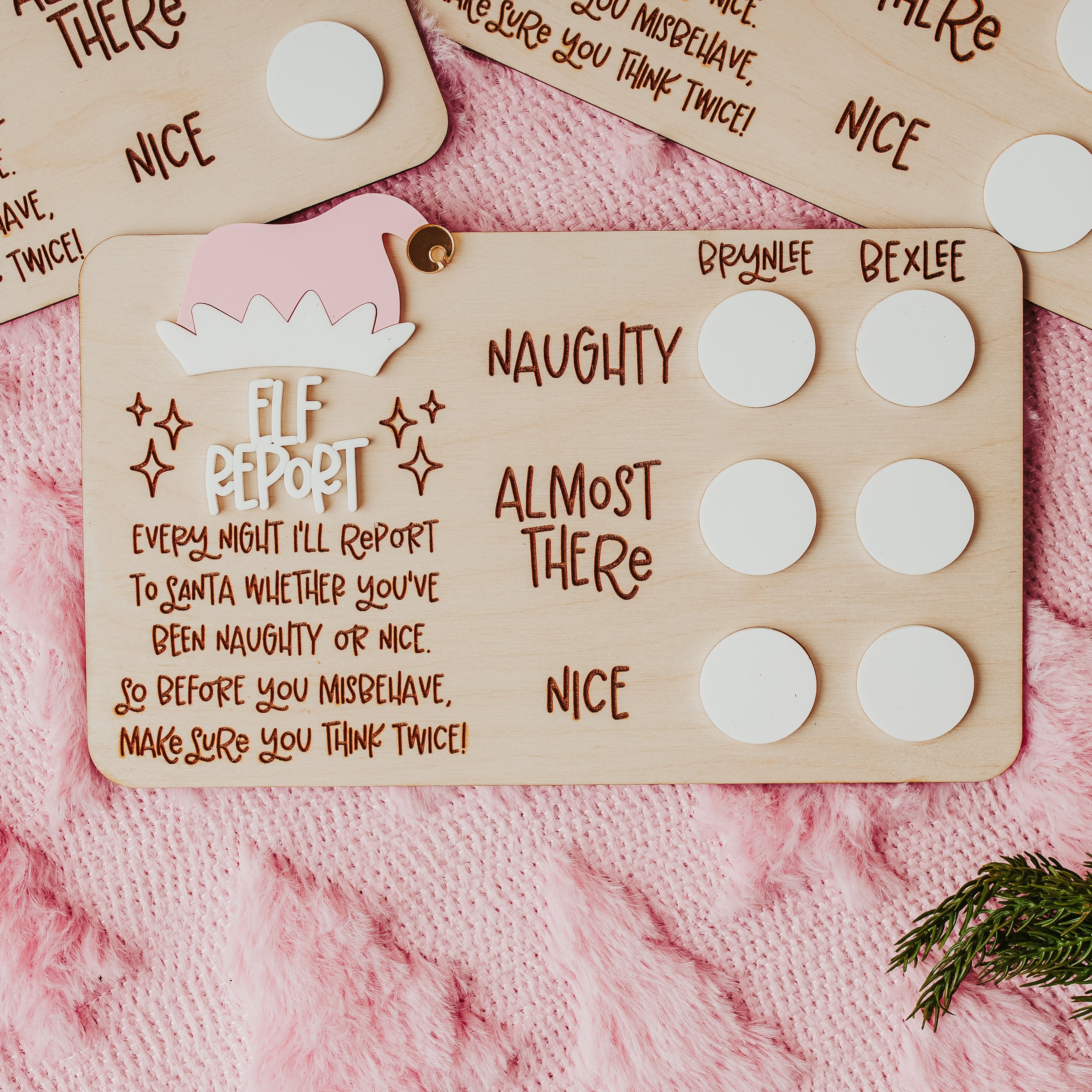 A close-up of a wooden Elf Report card with white circular white acrylic with personalized names, placed on a pink knit blanket with other matching cards partially visible.