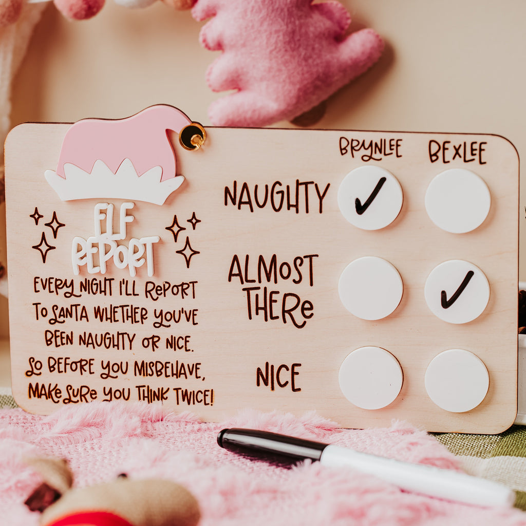 A wooden customized Elf Report checklist board with pink and white accents, displayed in a cozy, styled setting with plush toys and a pink blanket.