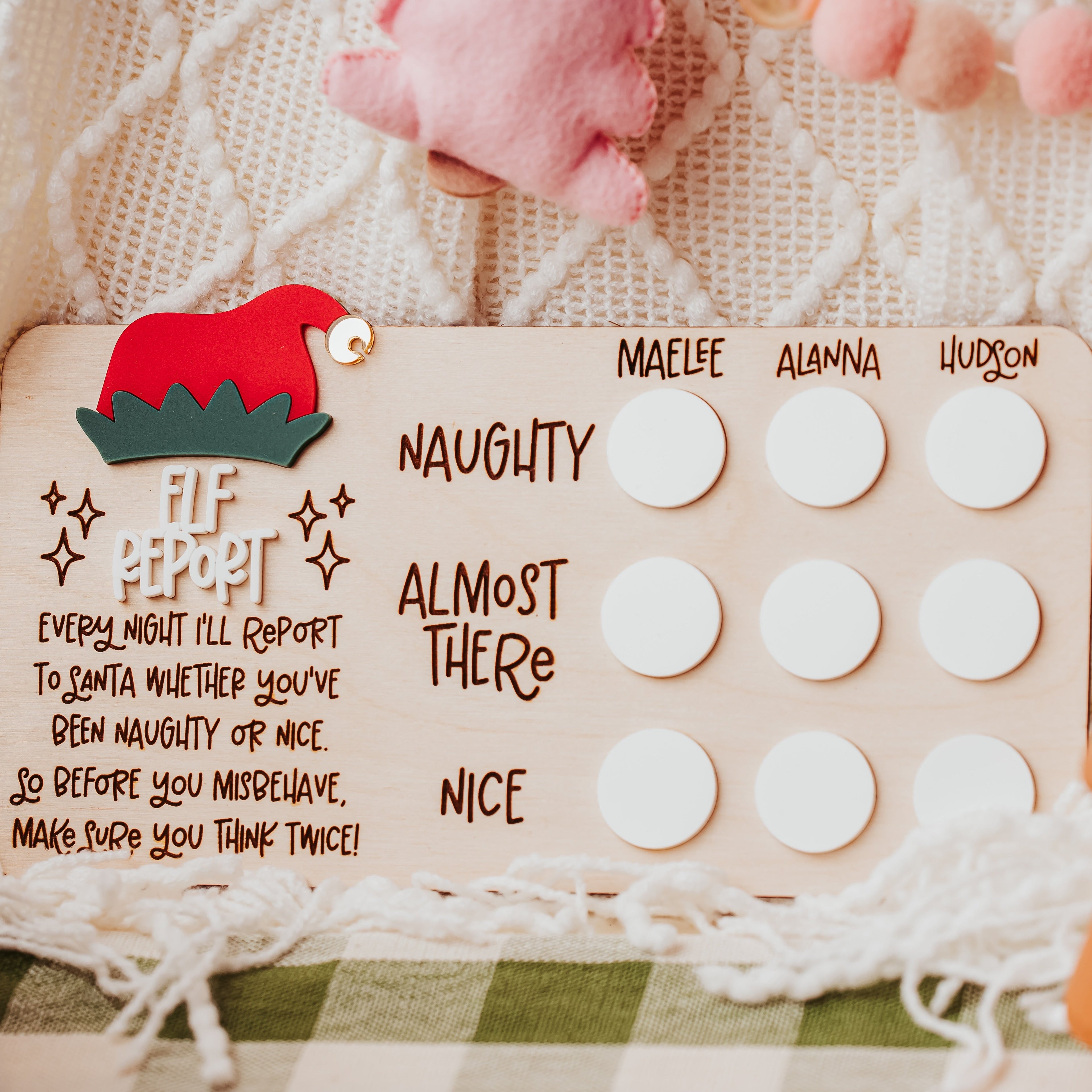 A festive Christmas elf report naughty or nice  board with a red and green elf  hat and white circular dry erase acrylic, styled on a knit blanket with soft pink and white pom-pom garlands.