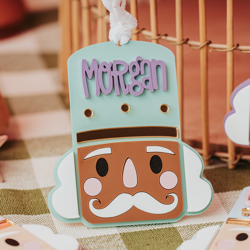 A close-up of a personalized nutcracker-shaped ornament with the name 'Morgan' in lavender lettering on a mint green top, displayed on a checkered tablecloth with other decorative ornaments in the background.