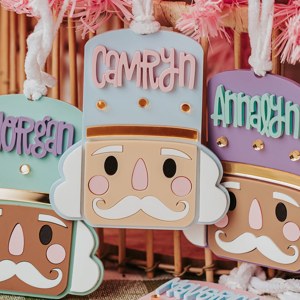 Personalized nutcracker ornaments hanging in a festive setting, featuring names like Camryn, Annalyn, Morgan, and Maverick on pastel-colored tags with white yarn hangers.