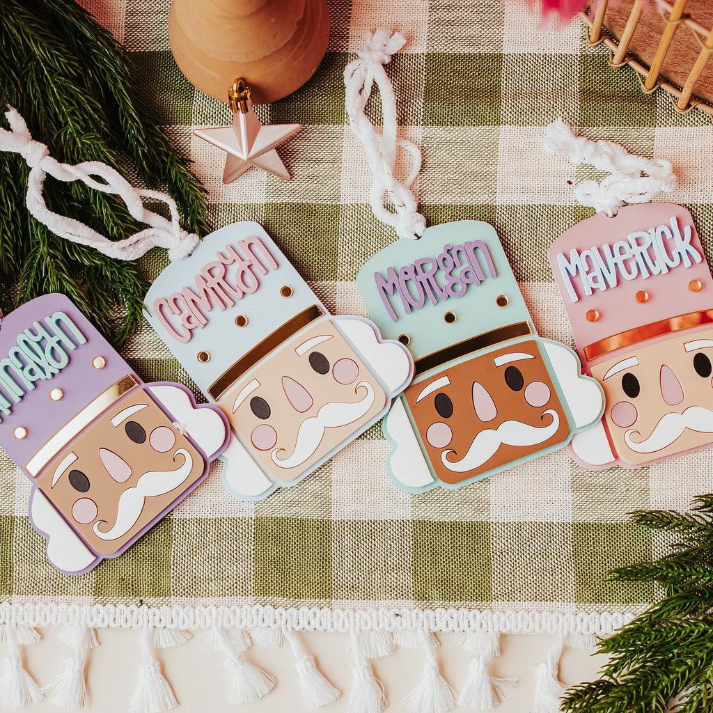 Four colorful nutcracker-shaped ornaments with names printed on them ('Annalyn', 'Camryn', 'Morgan', and 'MAVERICK'), arranged on a green and white checkered blanket with pine branches and holiday decor.