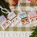 Four colorful nutcracker-shaped ornaments with names printed on them ('Annalyn', 'Camryn', 'Morgan', and 'MAVERICK'), arranged on a green and white checkered blanket with pine branches and holiday decor.