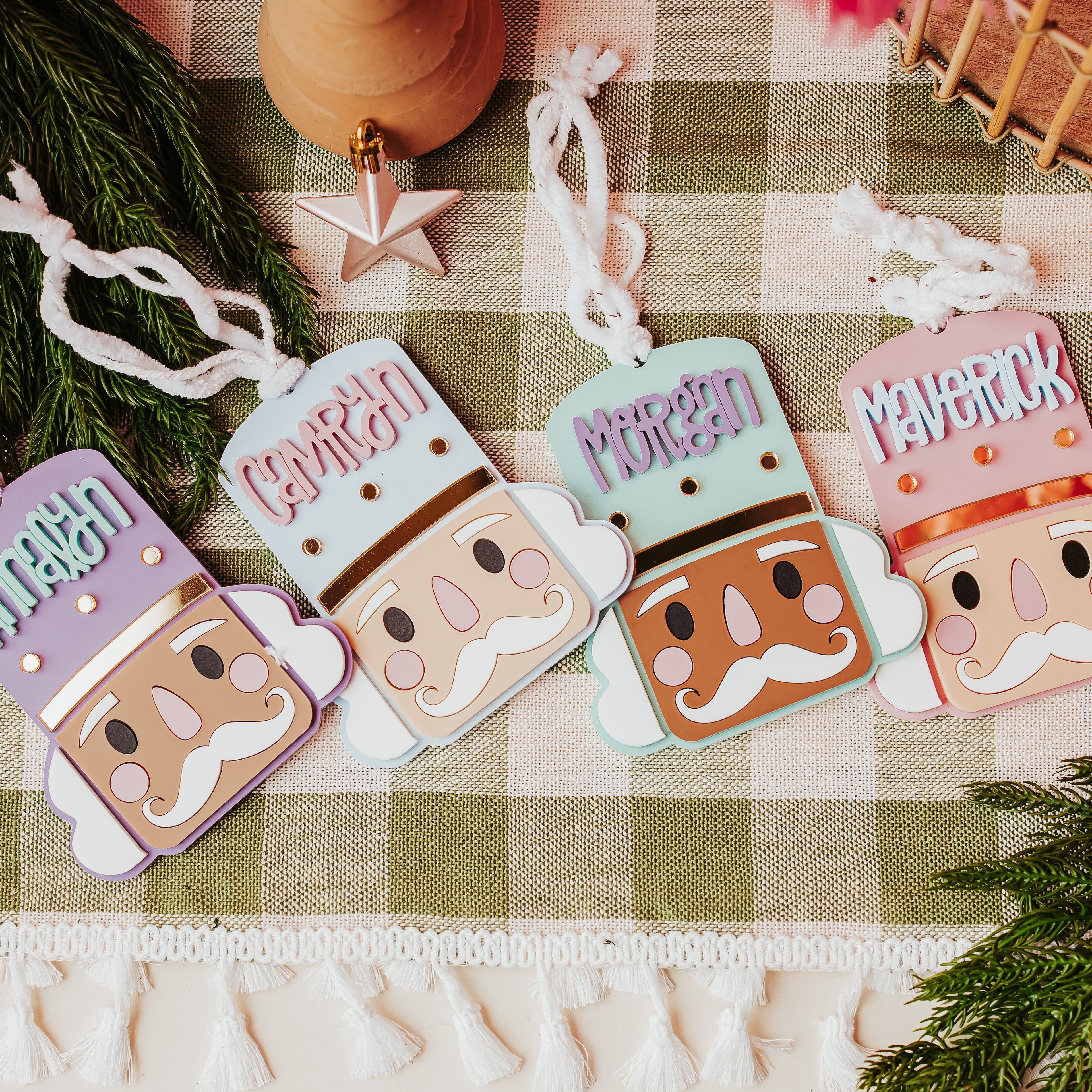 Four colorful nutcracker-shaped ornaments with names printed on them ('Annalyn', 'Camryn', 'Morgan', and 'MAVERICK'), arranged on a green and white checkered blanket with pine branches and holiday decor.
