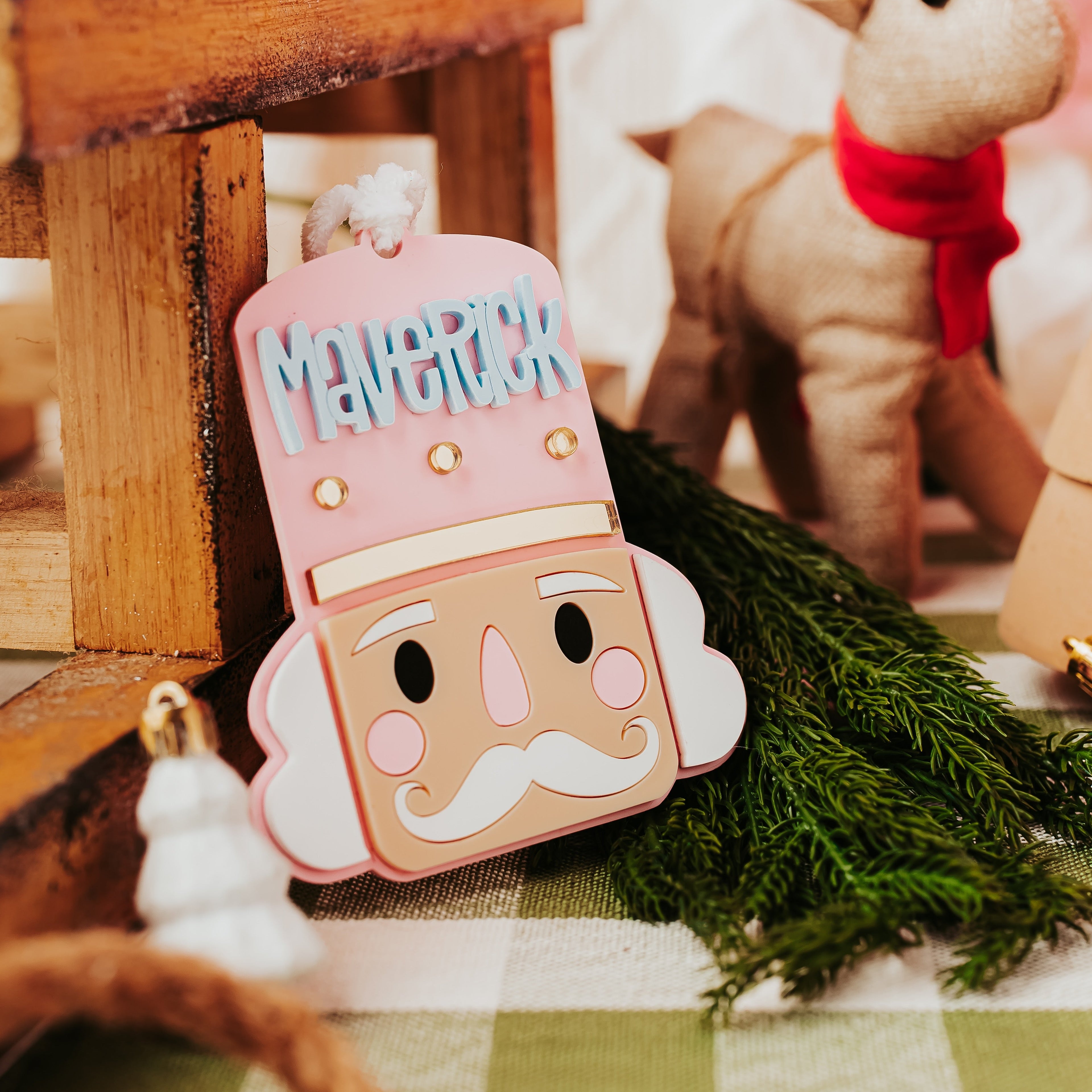 A close-up of a Christmas ornament shaped like a nutcracker face with the name 'MAVERICK' in blue letters on the pink hat. The ornament is styled on a green-and-white checkered surface with pine branches, a wooden crate, and a plush reindeer in the background.