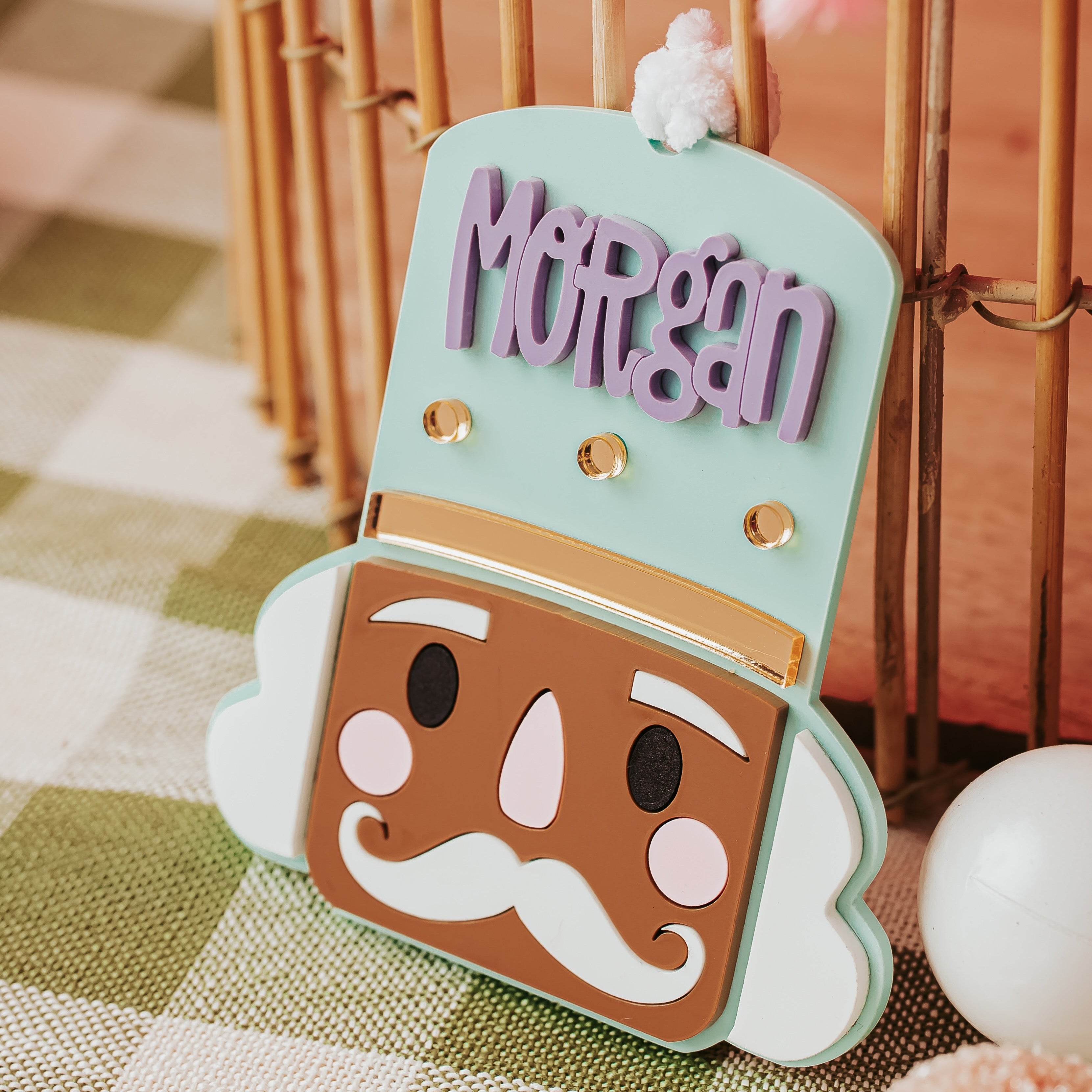 A personalized nutcracker-shaped ornament or sign with the name 'Morgan' in lavender letters, featuring a mint green top, brown face, white cheeks and mustache, and small gold dots, displayed on a green-and-white checkered tablecloth.