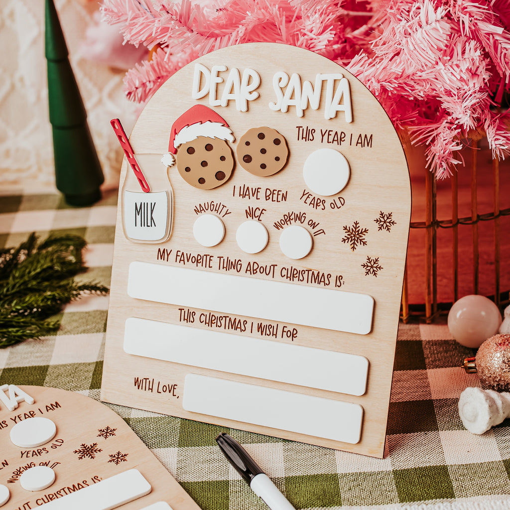 A wooden arch Christmas dry erase letter writing board for Santa, styled on a green and white checkered tablecloth with a pink artificial tree in the background.