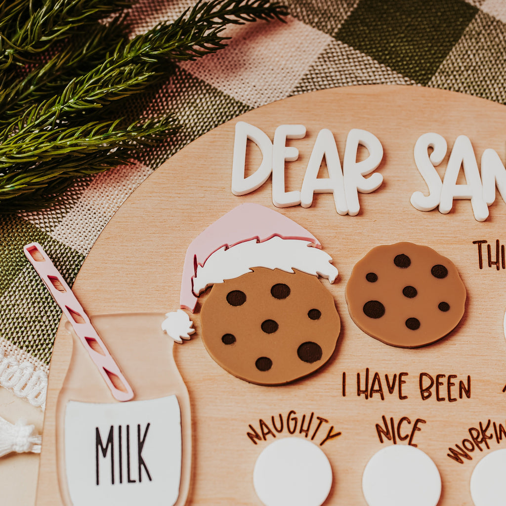 A festive wood arch Christmas letter to Santa dry erase letter. The kit includes white foam letters spelling 'DEAR SANTA', two brown cookie cutout shapes with black polka dots, a pink and white Santa hat, a milk cup with a red-and-white striped straw, and blank white oval stickers for personalization.