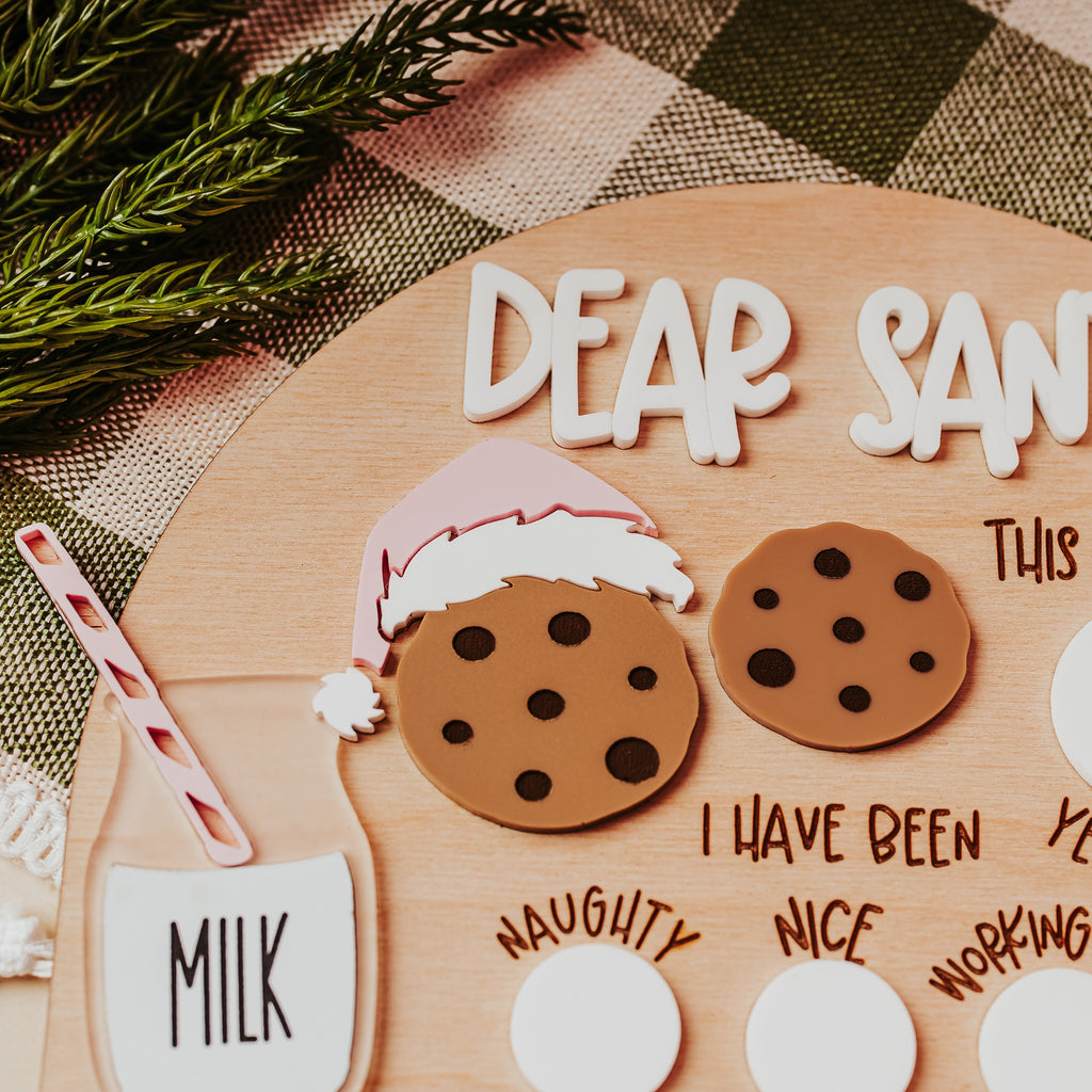 A festive handmade Christmas letter to Santa displayed on a wooden board. The kit includes white foam letters spelling 'DEAR SANTA', two brown cookie cutout shapes with black polka dots, a pink and white Santa hat, a milk cup with a red-and-white striped straw, and blank white oval stickers for personalization.