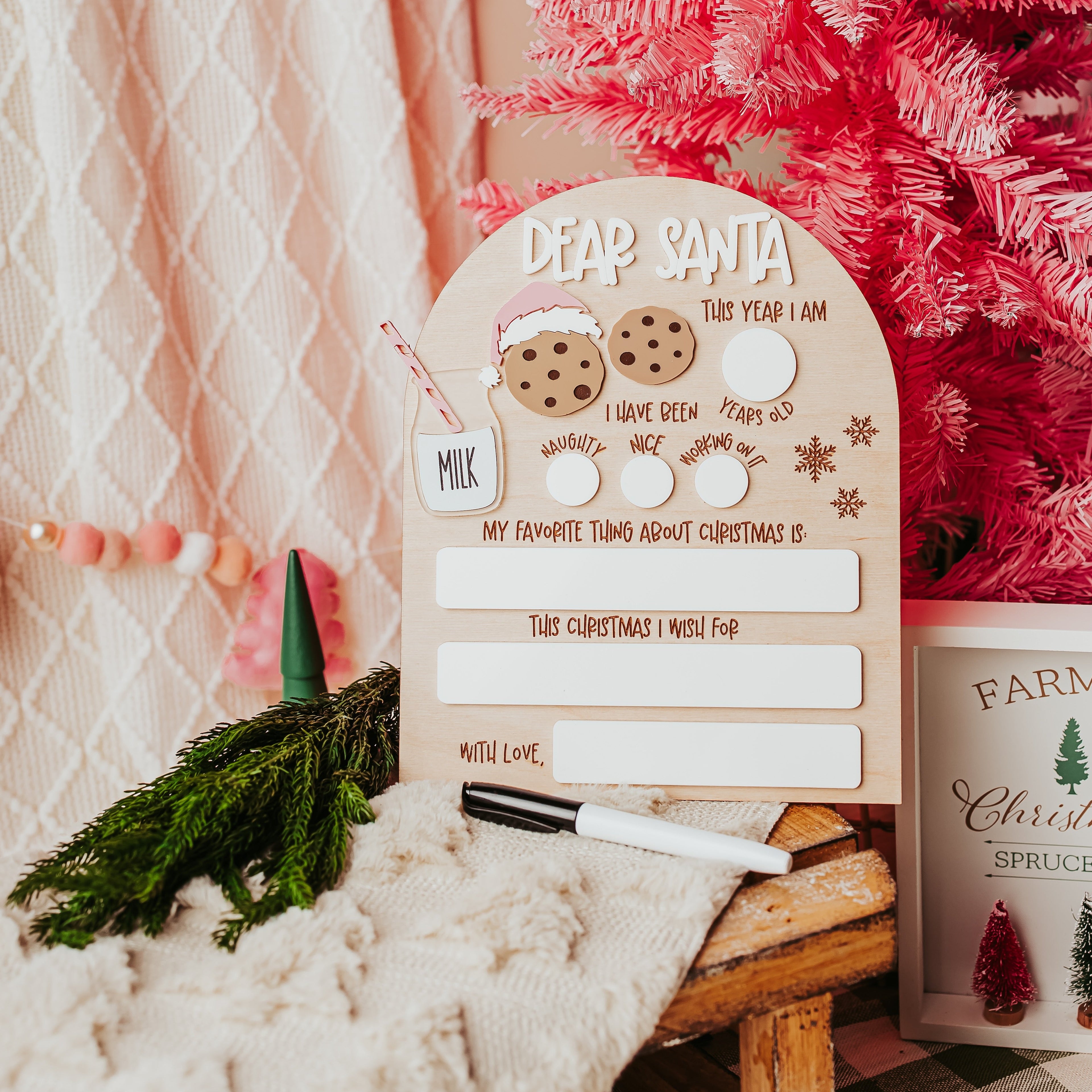A festive wooden arch holiday writing board set on a rustic wooden table next to a pink Christmas tree, featuring a beige arch-shaped board with 'DEAR SANTA' text, with white dry erase writing slots, cookie illustrations, and a black marker.