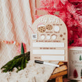 A festive holiday writing board set on a rustic wooden table next to a pink Christmas tree, featuring a beige arch-shaped board with 'DEAR SANTA' text, white writing slots, cookie illustrations, and a black marker.