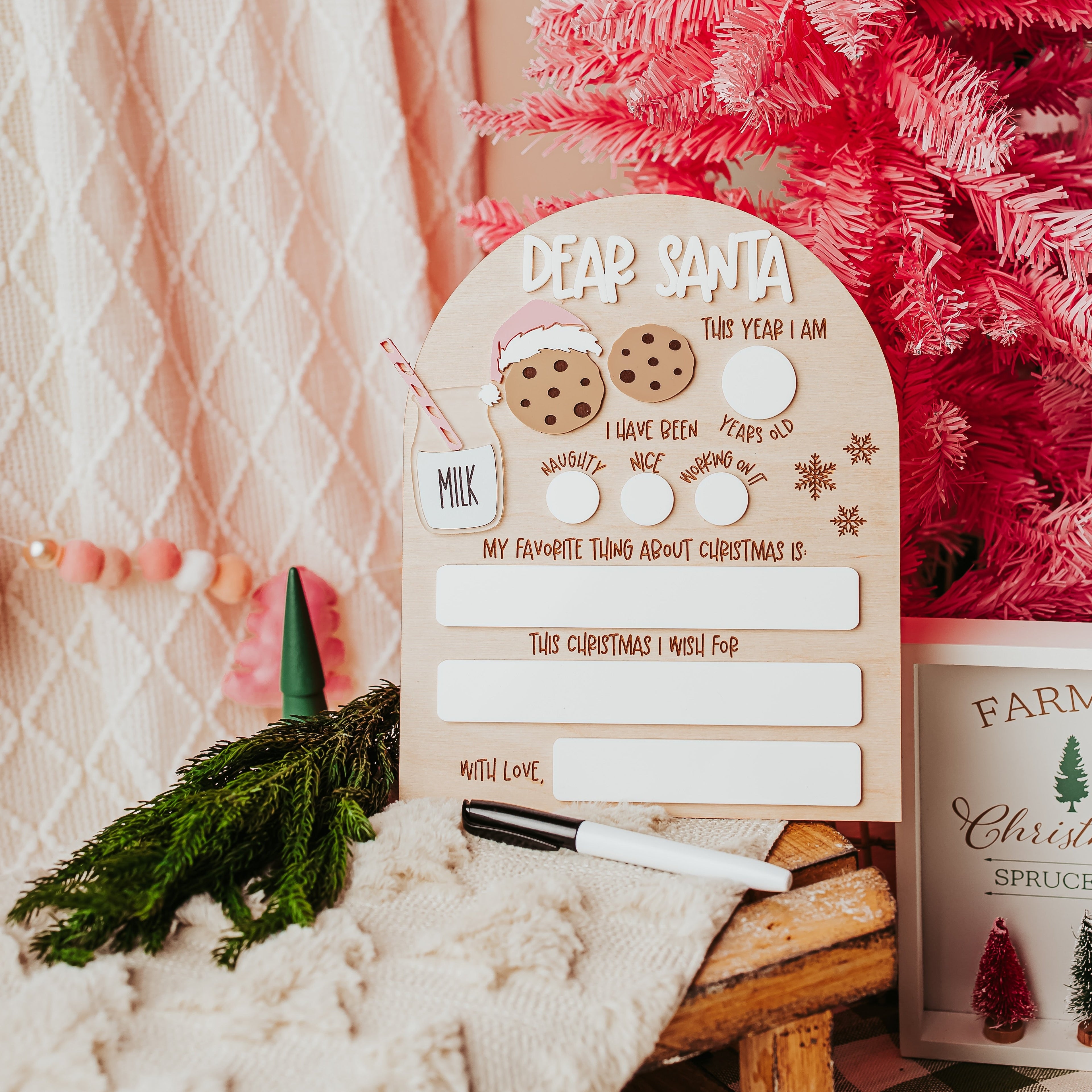 A festive holiday writing board set on a rustic wooden table next to a pink Christmas tree, featuring a beige arch-shaped board with 'DEAR SANTA' text, white writing slots, cookie illustrations, and a black marker.