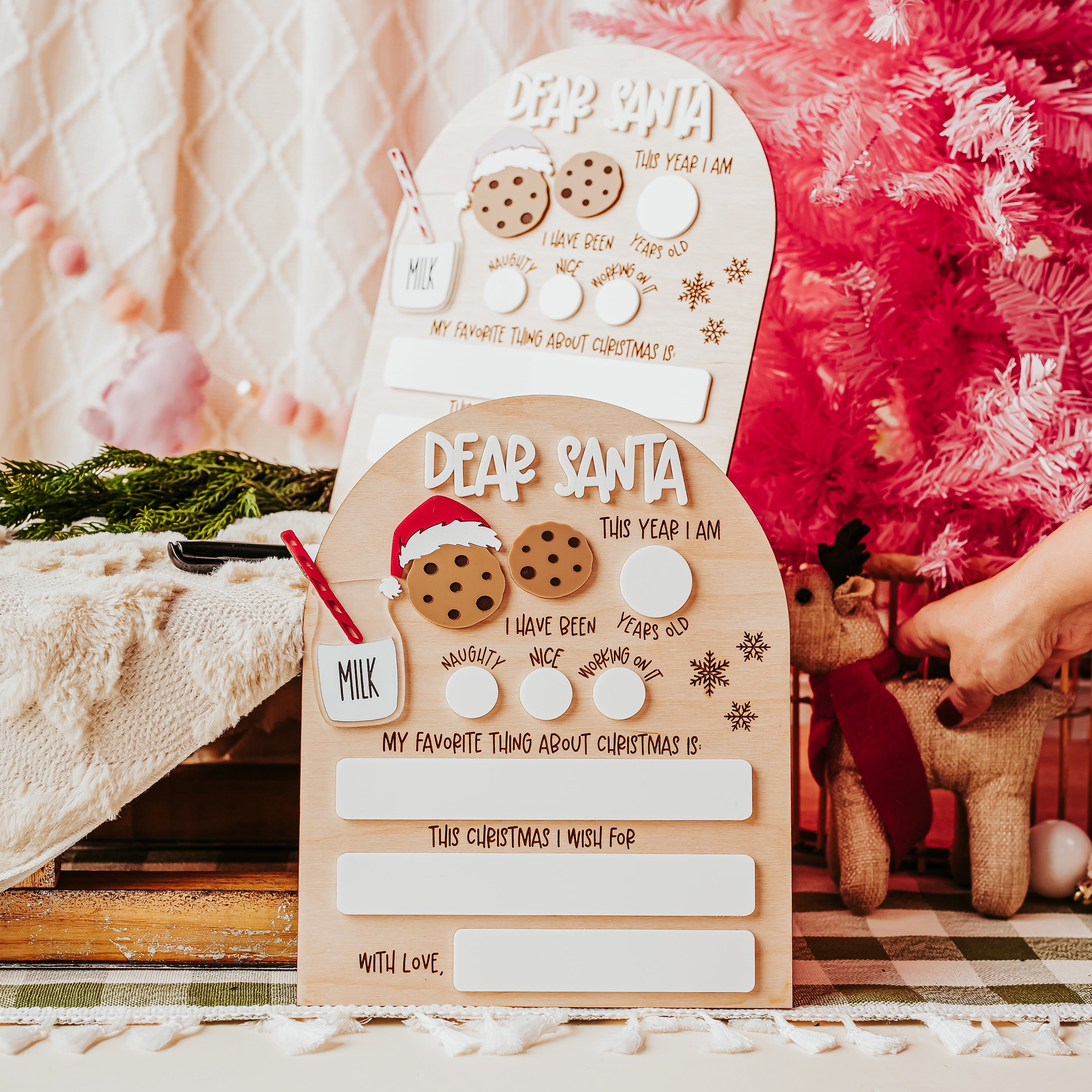 A Christmas dry erase "dear Santa' letter displayed on a table next to a pink Christmas tree, with a hand holding a wooden ornament nearby.