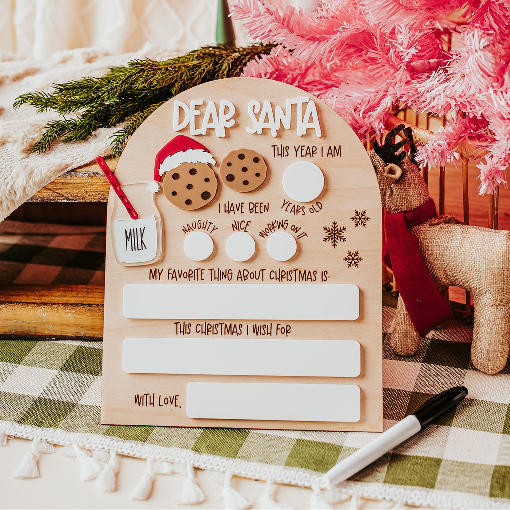 A decorative wooden arch Christmas dry erase 'Dear Santa' letter board styled on a green-and-white checkered tablecloth, surrounded by holiday props including a pink tree, a small reindeer, greenery, and a marker.