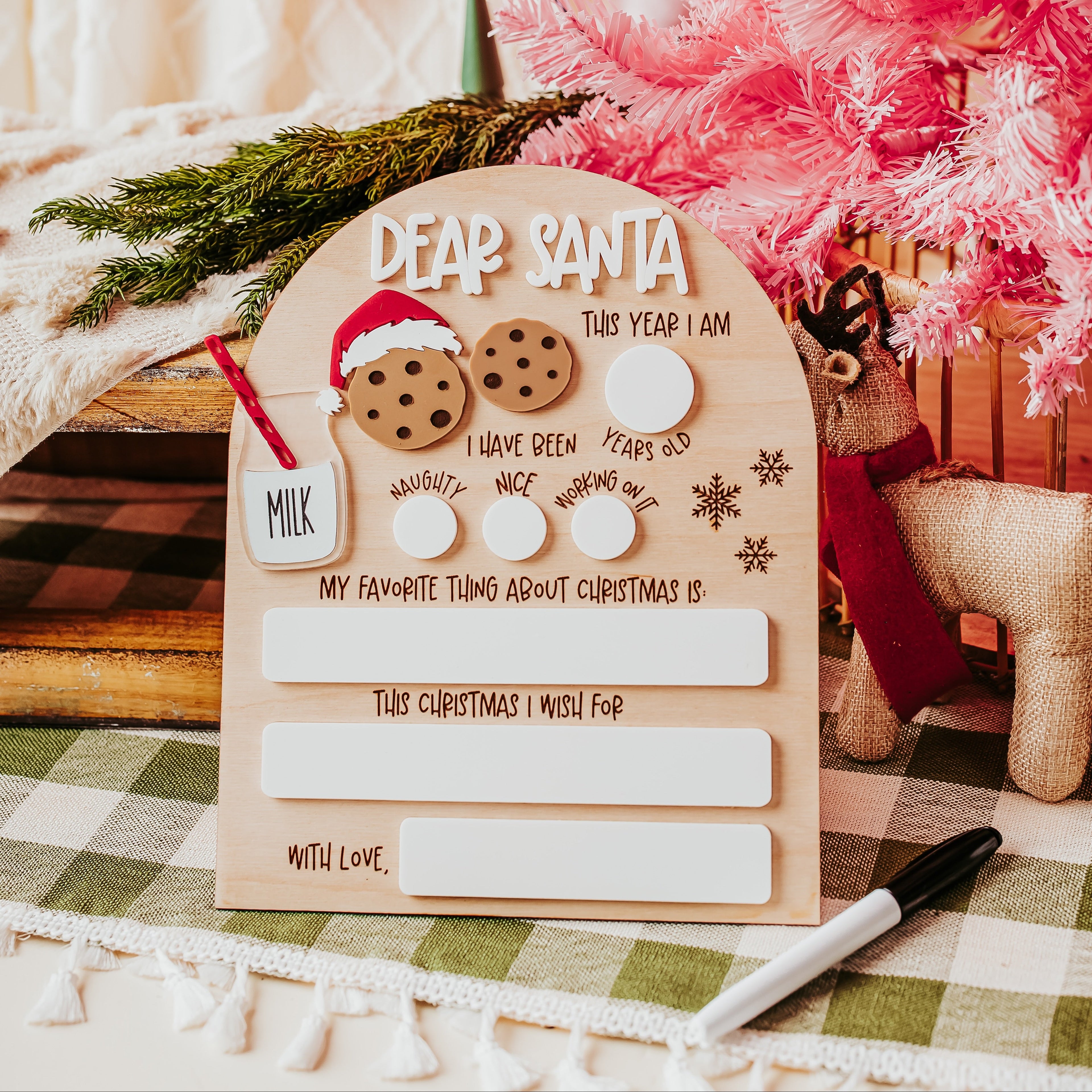 A decorative wooden arch Christmas dry erase 'Dear Santa' letter board styled on a green-and-white checkered tablecloth, surrounded by holiday props including a pink tree, a small reindeer, greenery, and a marker.
