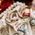 Three personalized wooden Christmas ornaments shaped like cars, displayed on a white tray with holiday decorations. The ornaments are engraved with names ('Elodie, 'Ledger', 'Carter'), messages ('My First Christmas', 'Merry Christmas'), the year '2025', and small colored car graphics with green trees on top.