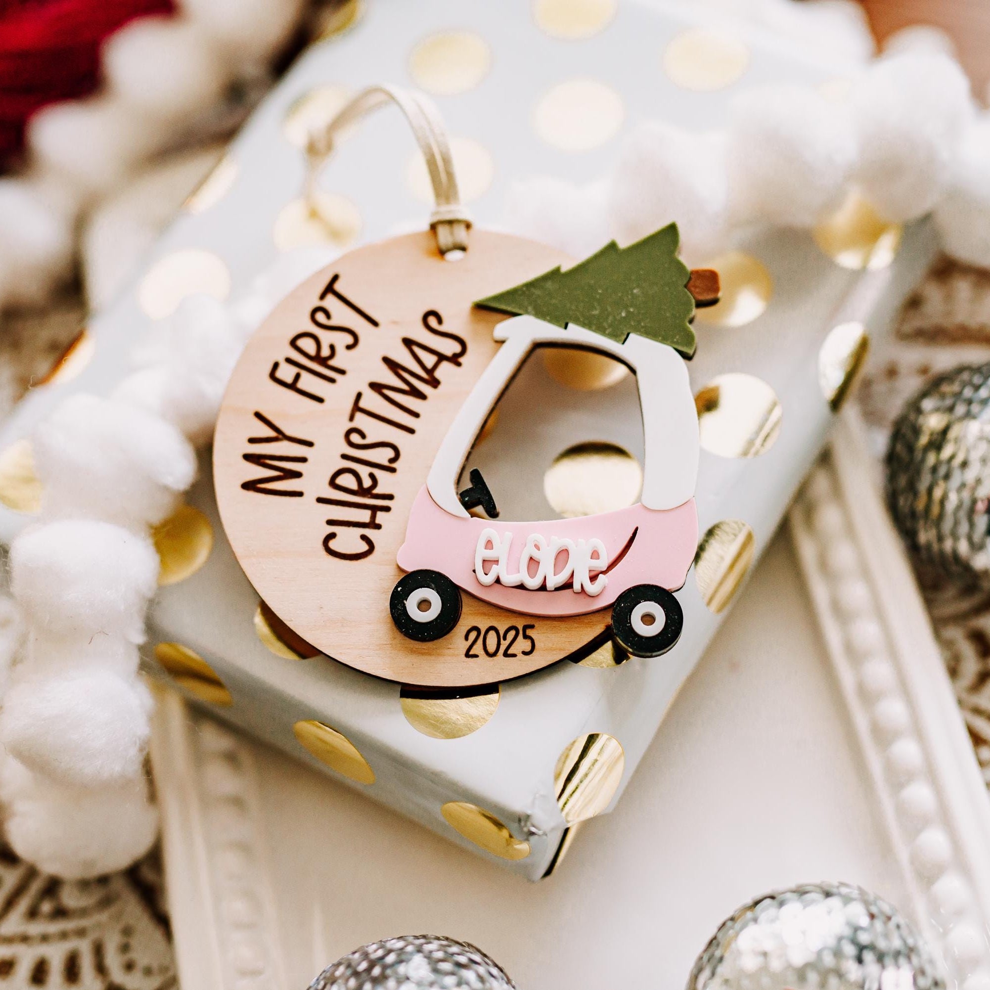 A close-up of a decorative Christmas ornament shaped like a pink cozy coupe car carrying a green tree, placed on top of a wrapped gift. The ornament has the words 'My First Christmas 2025' and the name 'Brielle' on it.