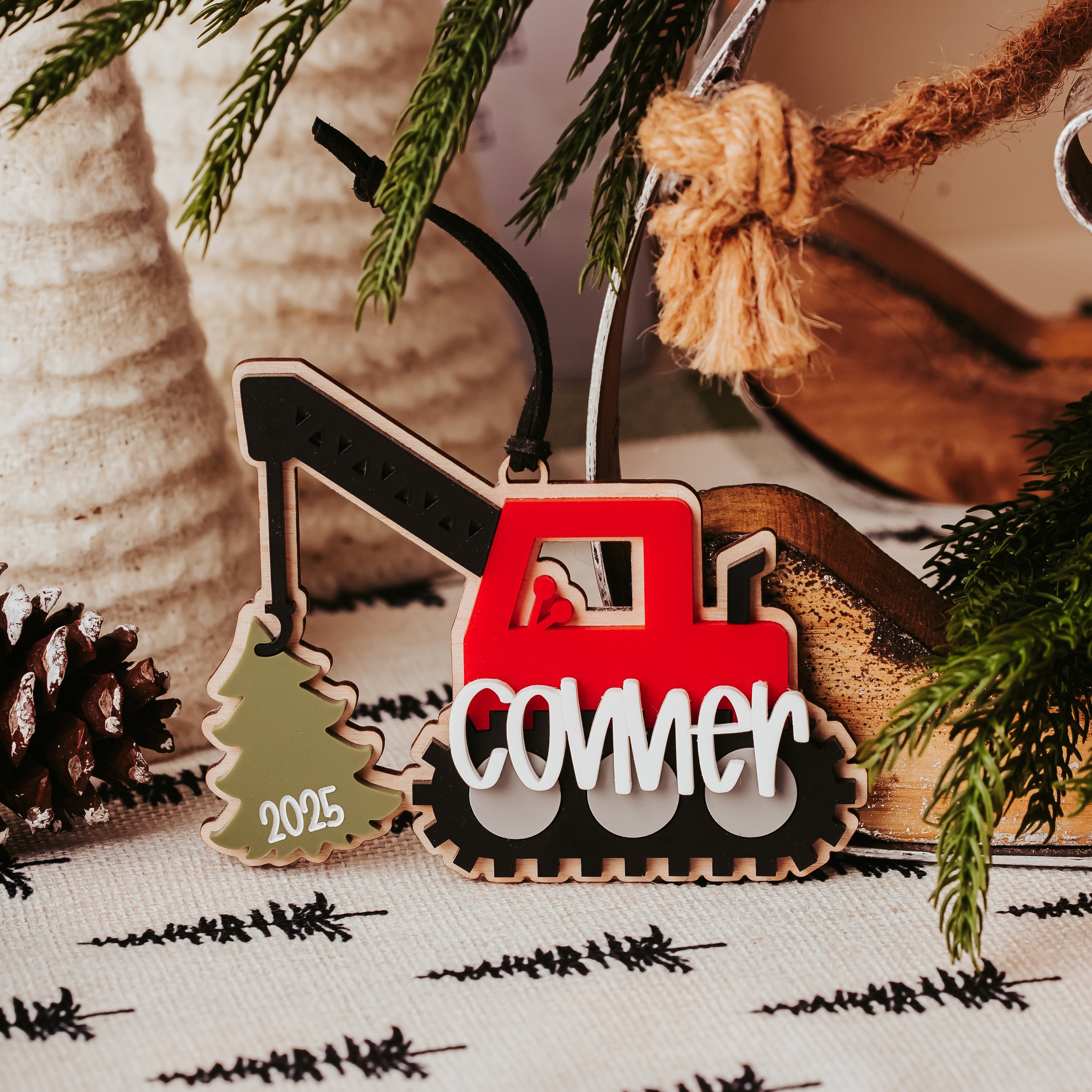 A close-up of a wood and acrylic Christmas ornament shaped like a red construction crane with the word 'conner' in white script, hanging near evergreen branches and a pinecone on a patterned fabric surface.