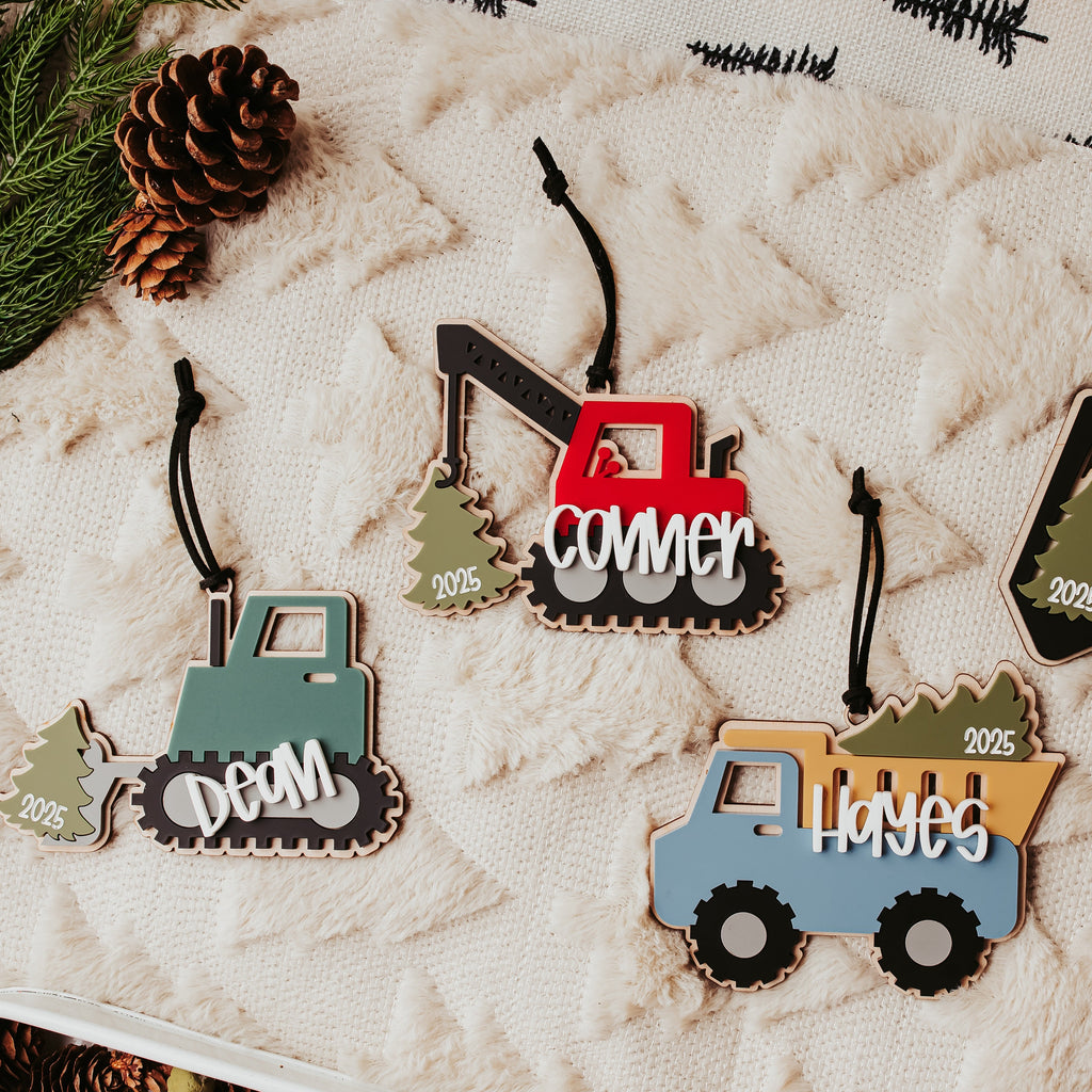 Four personalized wood and acrylic construction vehicle ornaments hang from black strings on a textured cream quilt. The ornaments include a pine green bulldozer with the name 'DEAN', a red crane with 'conner', and a blue dump truck with 'HAYES'.