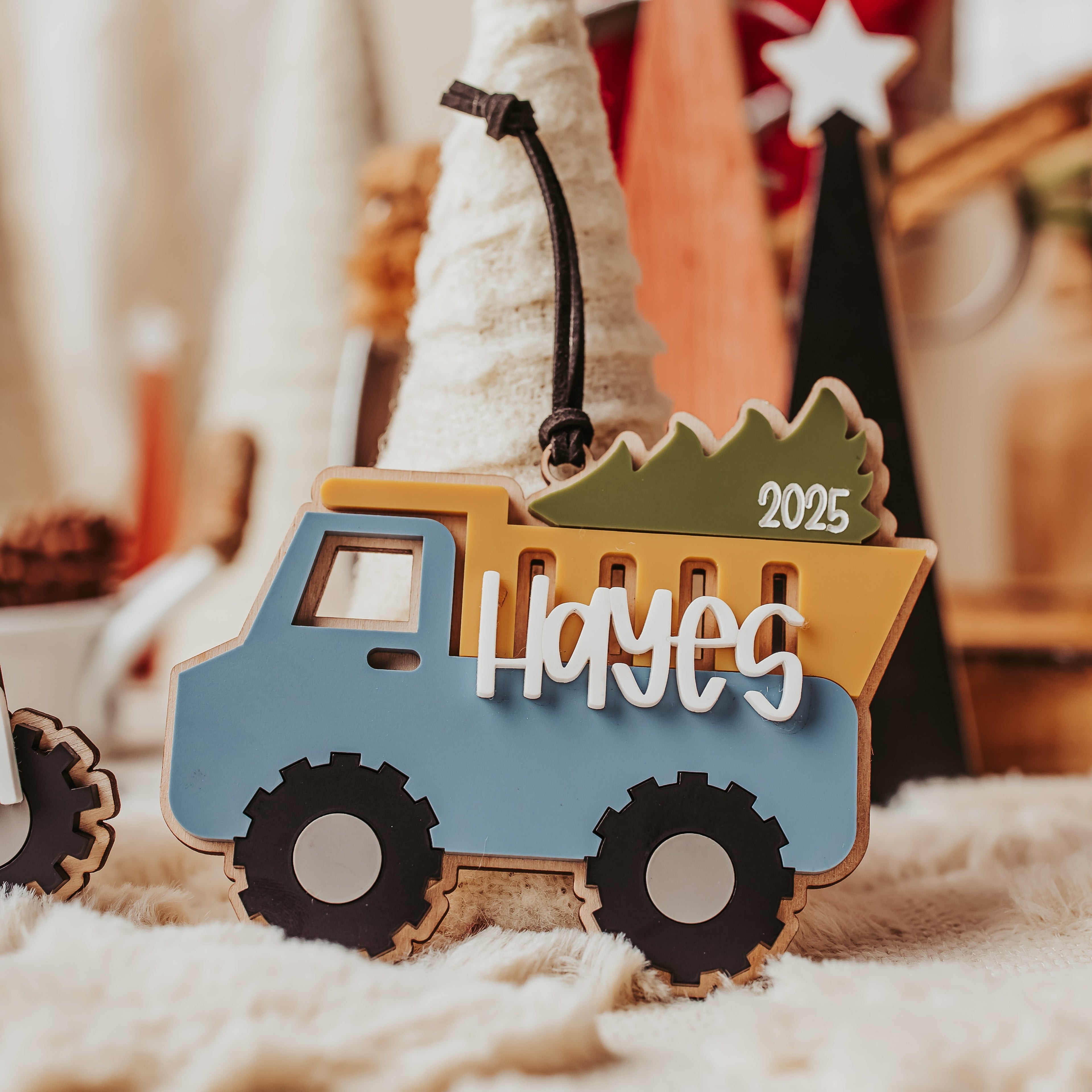 A personalized wood and acrylic Christmas ornament shaped like a dump truck carrying a Christmas tree, with the name 'Hayes' and the year '2025' cut into the side. The ornament is styled on a soft cream blanket with a blurred festive background.