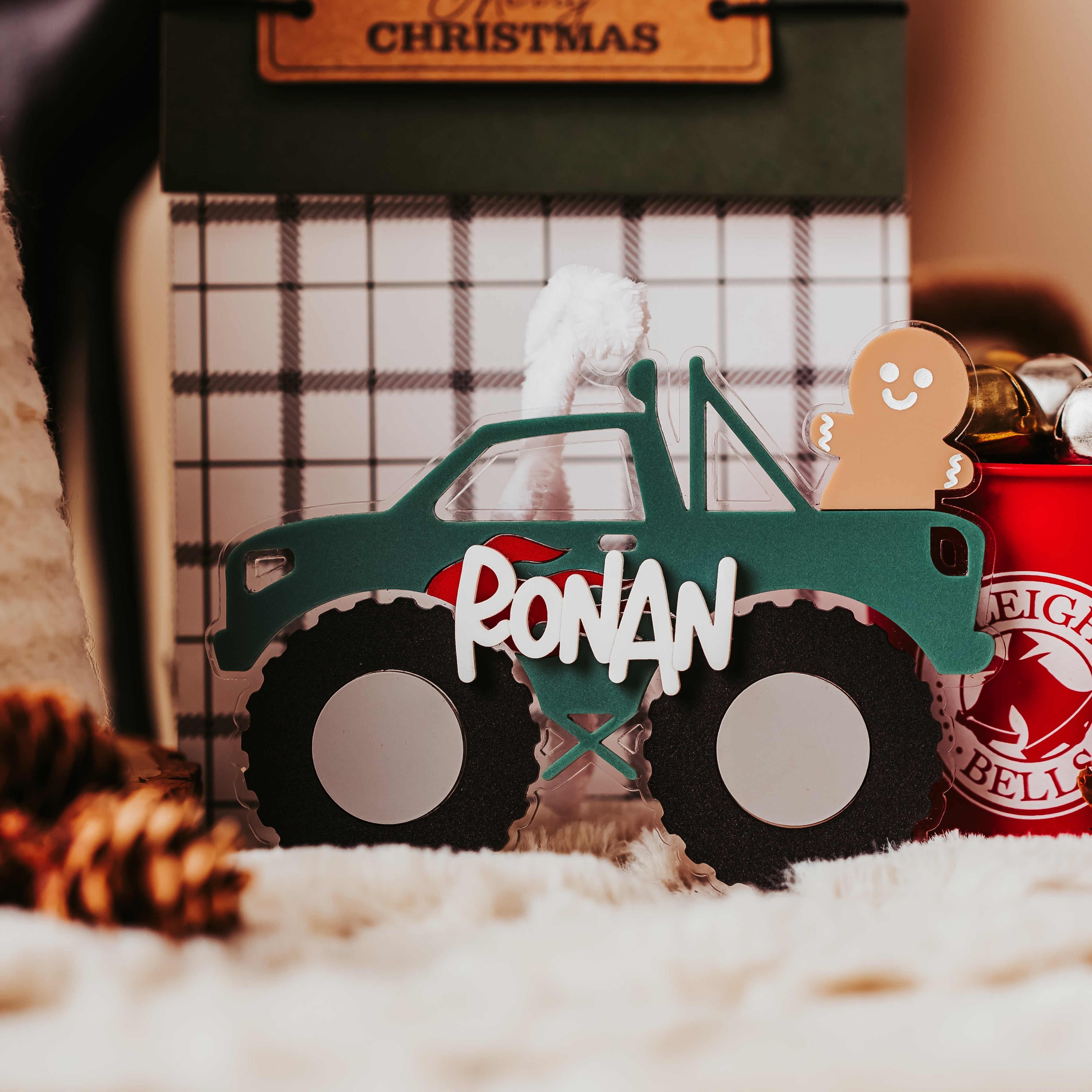 A personalized Christmas monster truck ornament with the name 'RONAN' in white across the front, styled on a white textured surface with holiday props including a gingerbread cookie, pinecone, and a 'Merry Christmas' card in the background.