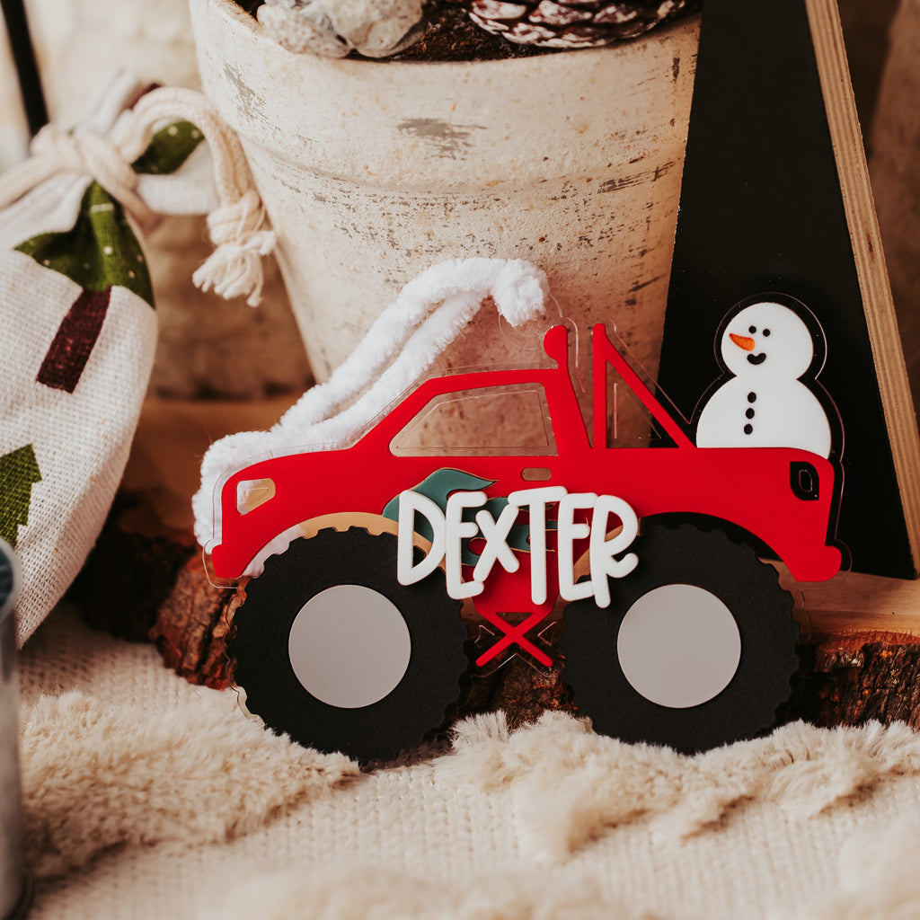 A personalized red monster truck  ornament with the name 'DEXTER' in white on the side, styled with pinecones in a birch bark pot and a Christmas tree decoration on a cozy, rustic background.